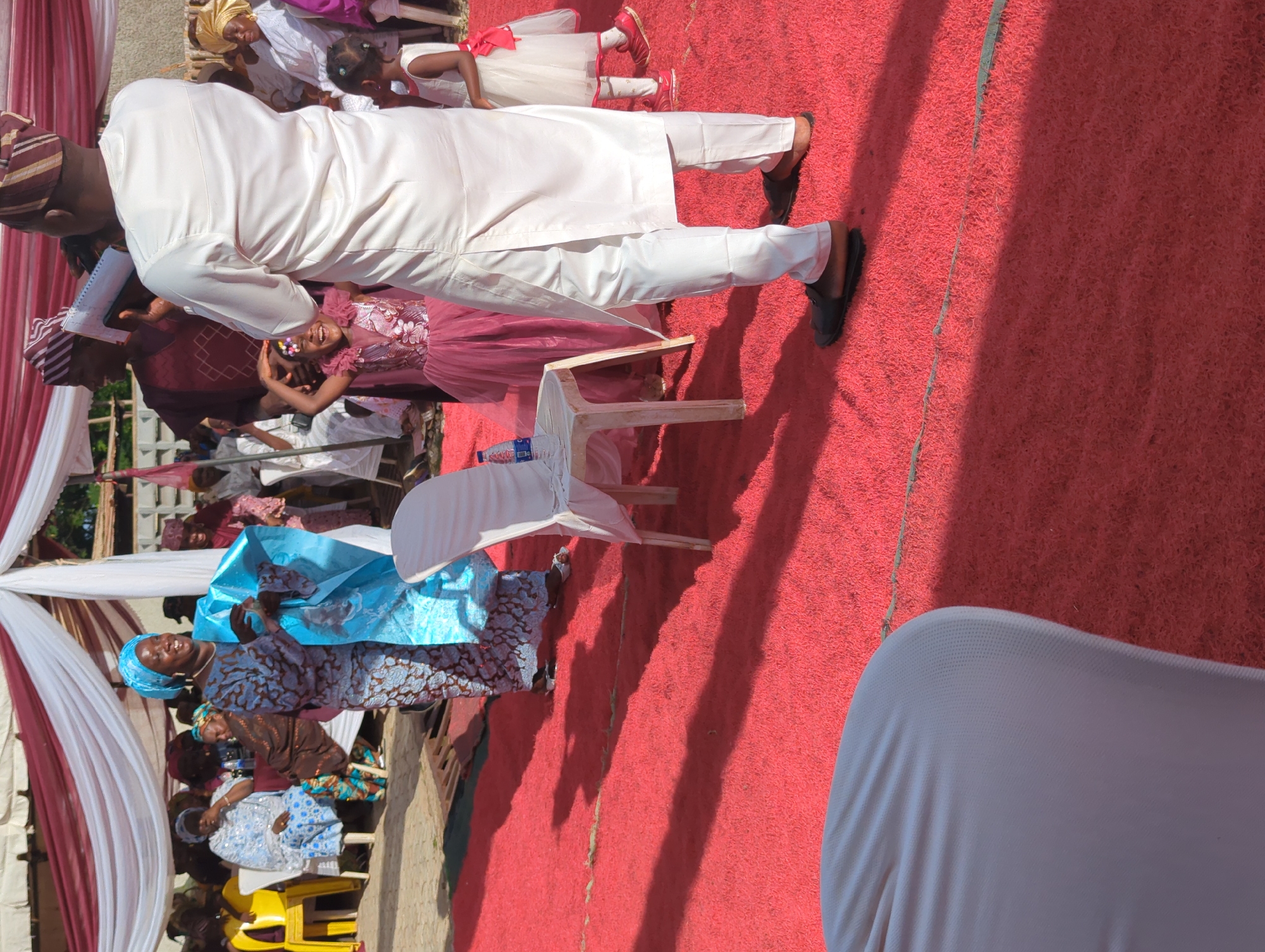 The image captures an outdoor event in Kabala, Nigeria, beneath a white and red striped canopy. A vibrant red, textured carpet covers the ground, with prominent shadows indicating strong sunlight.
In the center-left, a woman wearing a blue patterned traditional Nigerian dress and a matching headwrap smiles widely while holding a piece of light blue fabric. Adjacent to her, a white plastic chair holds a clear plastic bottle of water. To the right of the chair, a young girl in a sleeveless pink gown with floral embellishments and colorful hair accessories stands, looking up and smiling exuberantly with her hands near her head. Standing with his back to the viewer, a man in a white traditional Nigerian tunic and trousers, paired with a striped brown and white cap, holds a notebook or small device in his hands. He faces the young girl and other attendees. To his right, a smaller girl in a white dress with a red sash and red shoes stands still.
In the background, under the canopy, multiple individuals are seated on a combination of yellow plastic chairs and white fabric-covered chairs, observing the event. The man in white appears to be addressing or leading a portion of the gathering.