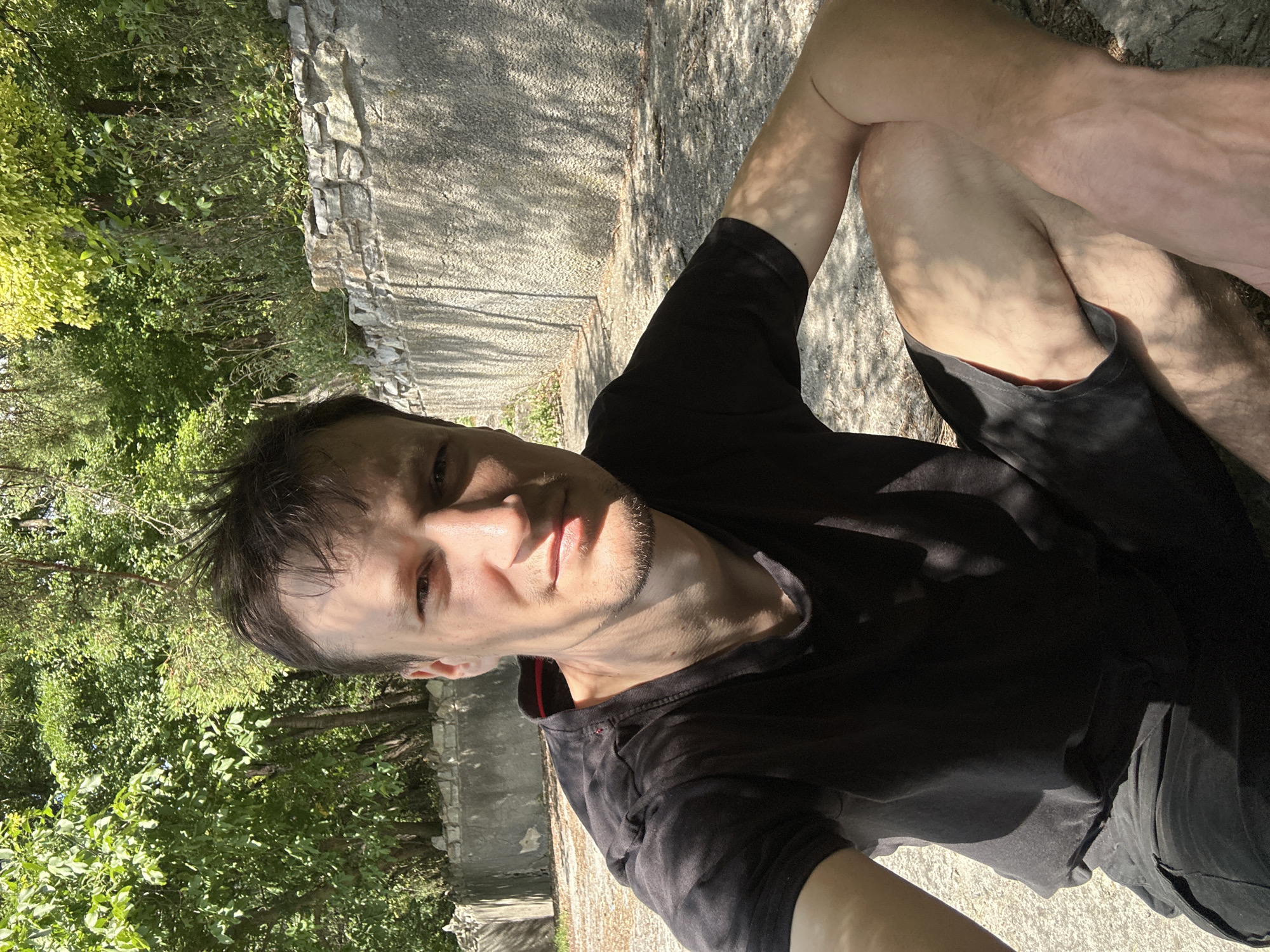 A man is sitting on the ground, possibly in the city of Makiv, Ukraine, in a sun-dappled area with trees and a stone wall behind him. He is wearing a black t-shirt and black shorts. The man is looking directly at the camera, and the lighting suggests it is daytime, with sunlight casting shadows. The background features lush green foliage and a textured stone wall.