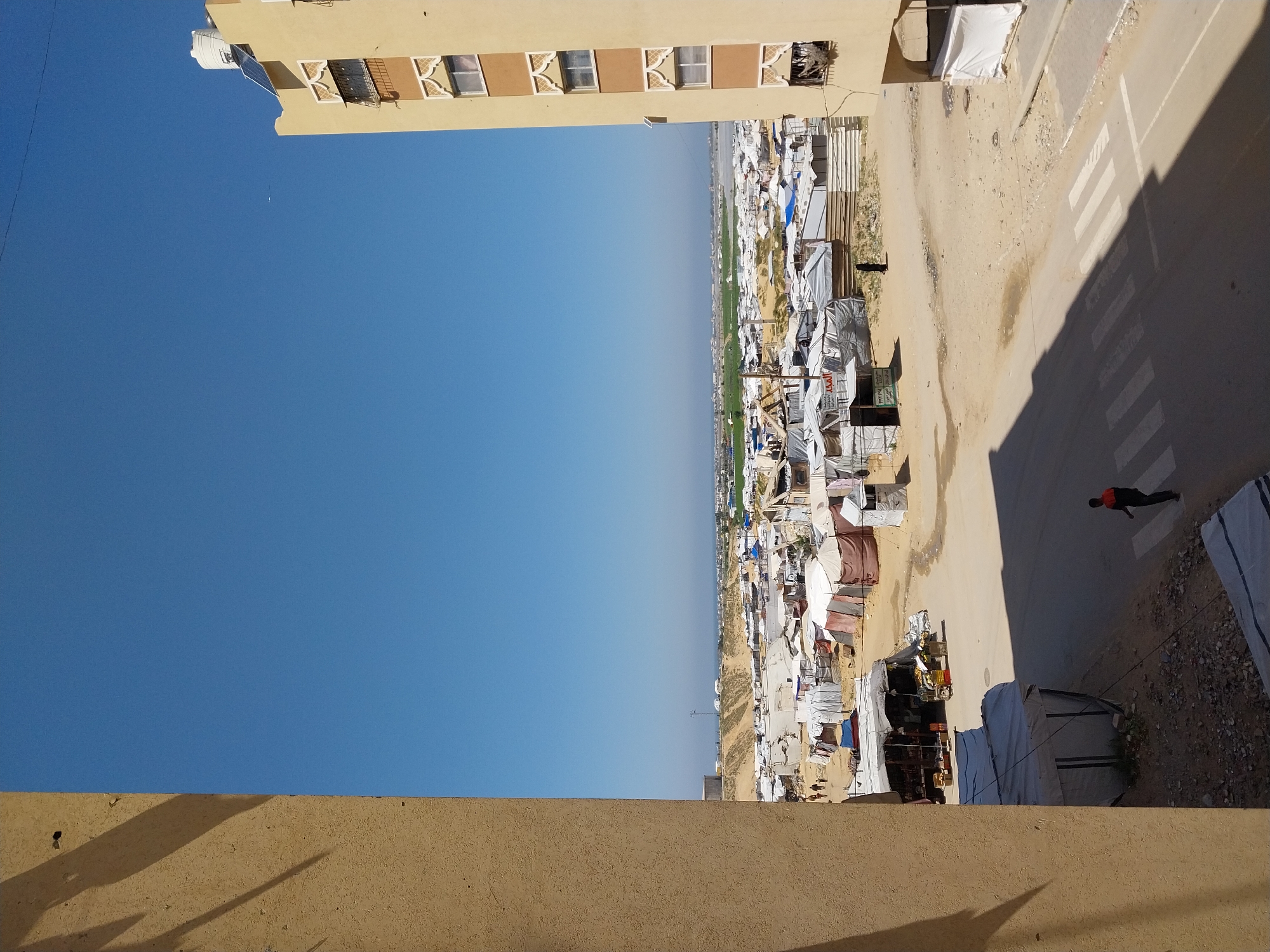 The image depicts a partial view of what appears to be a refugee camp or informal settlement located in the city of Unknown, Unknown. The scene is captured from an elevated position, possibly from a nearby building.
In the background, there is a clear, blue sky. The camp consists of numerous makeshift shelters constructed from a variety of materials including cloth, tarps, and corrugated metal. There is also vegetation visible amidst the shelters. Adjacent to the camp is a paved road with a pedestrian crosswalk. A person wearing a red shirt is walking across the crosswalk.
Parts of two buildings are also visible in the frame. The building at the top has multiple windows, while the second building, at the bottom of the frame, is closer in proximity to the viewer, and is a solid structure.