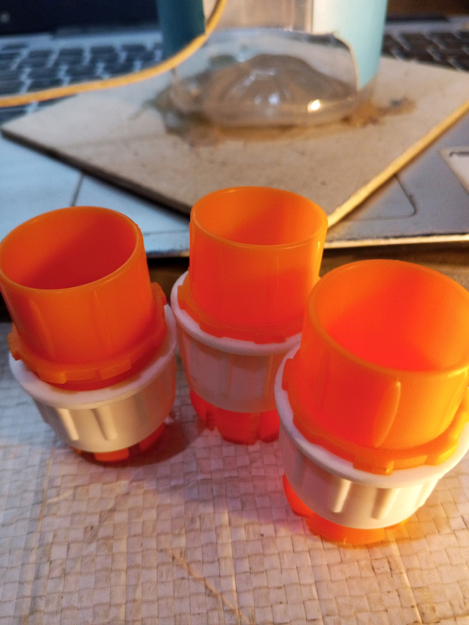 Three orange and white plastic caps are arranged in a triangular formation on a woven surface. The caps are cylindrical with a wider, flared top and a narrower base. They appear to be for bottles or containers. In the blurred background, a laptop keyboard and a light blue object are visible. The image is taken from a slightly elevated angle. The location is not identifiable within the city of Karawang, Indonesia, based on the visual information provided.