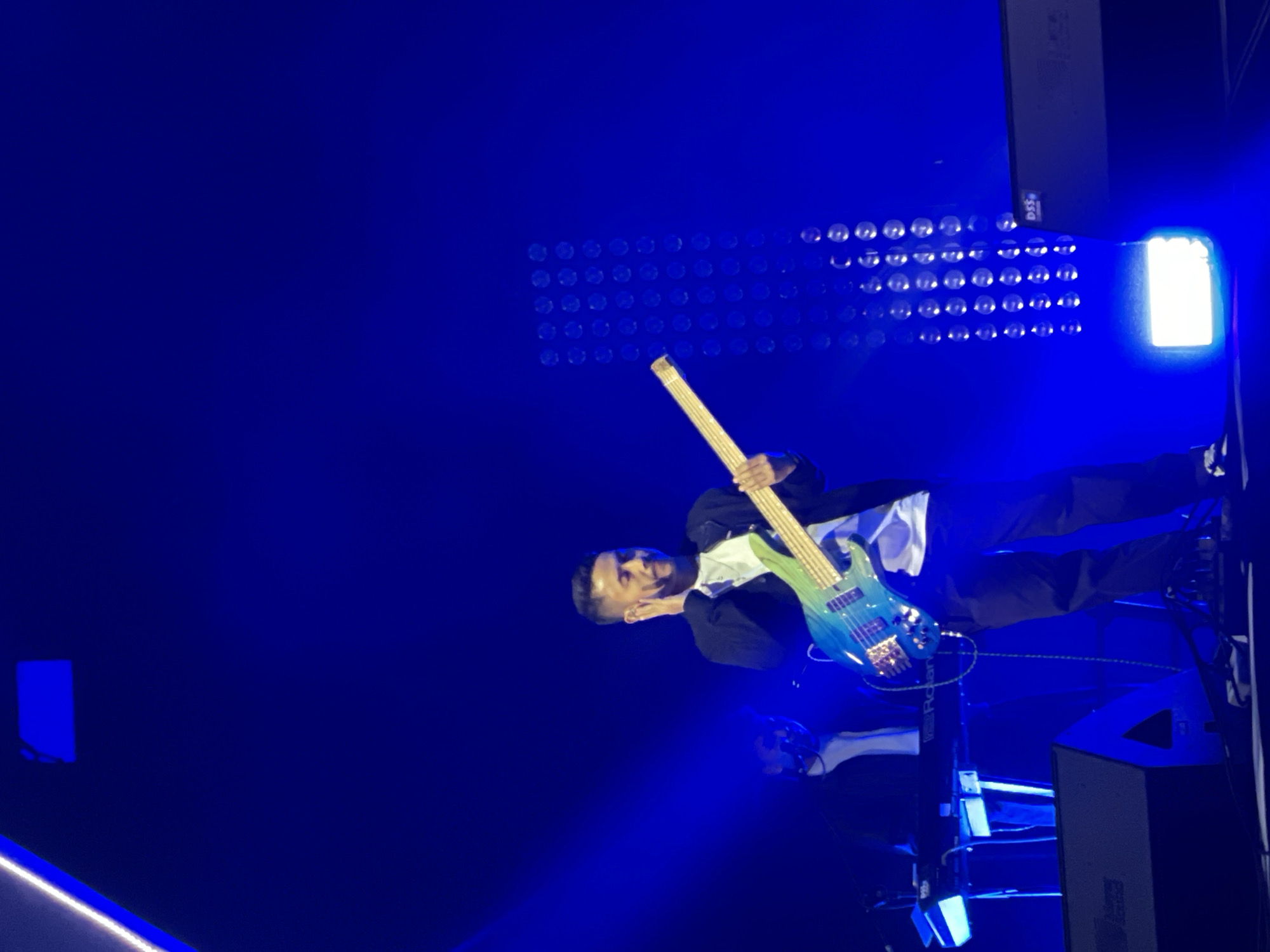 The image shows a musician playing a bass guitar on a stage bathed in blue light. A grid of lights and a speaker stack are visible in the background. The location, Salembaran, Indonesia, is not apparent in the image.