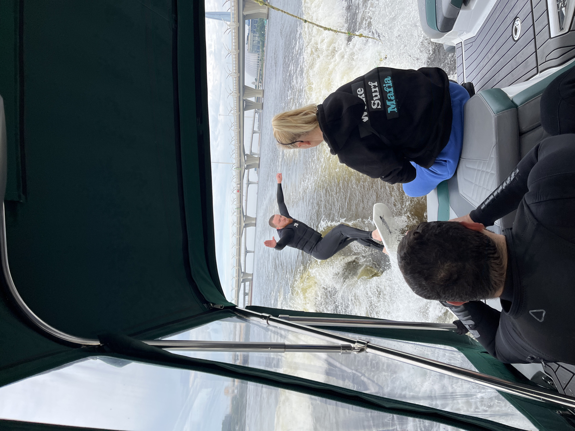 The image depicts a wake surfing activity on the Neva River in Saint Petersburg, Russia. In the foreground, two individuals are seated in a boat, facing away from the camera and towards the action. The person on the right is wearing a black hoodie with "Wake Surf Mafia" printed on the back in white and teal lettering. The person on the left is wearing a black wetsuit and is partially visible from the back of their head.
In the mid-ground, a male surfer is actively wake surfing on a white board. He is wearing a black wetsuit with a life vest over it, and his body is positioned in a dynamic stance, riding the wake generated by the boat.
The background shows a wide expanse of the Neva River with choppy water, indicating the boat's movement. Dominating the background is a large, modern bridge structure, likely part of the Western High-Speed Diameter, a major highway in Saint Petersburg, spanning the river. The distinctive architecture of the Lakhta Center, a skyscraper, is also visible in the distance to the right, further confirming the location. The sky is overcast with a muted, grey tone. The overall scene captures a recreational activity set against a prominent urban backdrop.