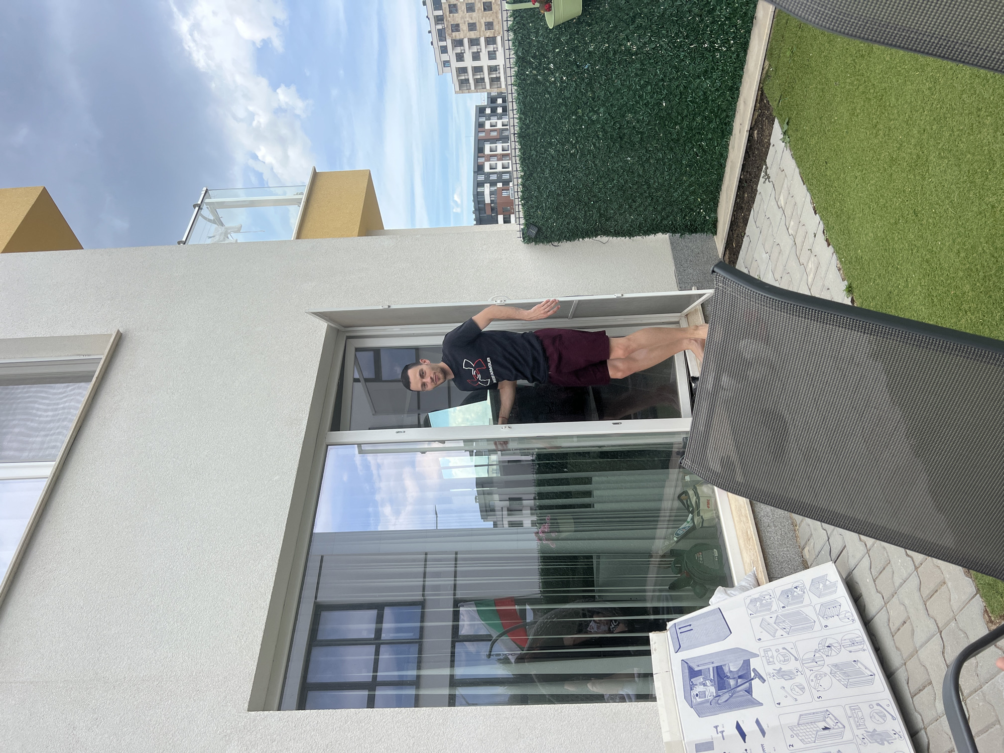 A man stands on a patio or balcony, seemingly doing a handstand against a large glass sliding door. He is wearing a dark t-shirt with a graphic design and maroon shorts. The door reflects the sky and buildings in the background. The setting appears to be a modern building with white walls and yellow accents. Beyond the patio, there's a patch of artificial turf and a wall covered in greenery, suggesting an urban or suburban outdoor space. The sky is cloudy and bright, indicating it's daytime. A box with assembly instructions is visible in the foreground, hinting at a recent purchase or project. The man's facial expression is neutral, and the overall mood is casual and active.