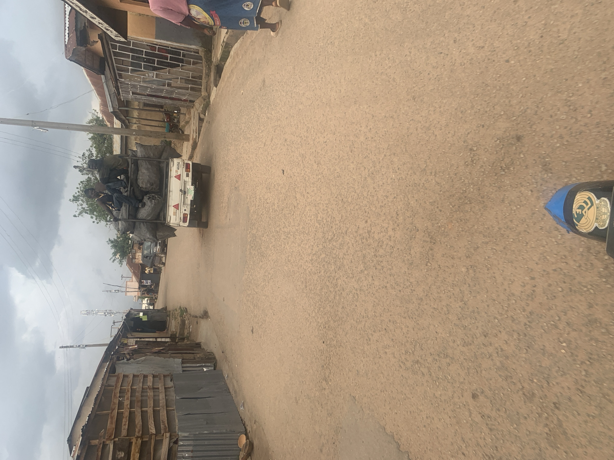 The image depicts a street scene in Gura Top, Nigeria, under an overcast sky. The perspective suggests the photo was taken from a moving vehicle, likely a motorcycle, as evidenced by the blue and black object in the foreground that appears to be part of the vehicle. The street is paved with asphalt, and the buildings lining both sides appear to be simple, tin-roofed structures.
On the left side of the street, a structure made of wooden planks and corrugated metal is visible. Several round, shallow bowls are placed on the ground in front of it.
On the right side, a white pickup truck is the central focus of the scene. It is heavily laden with large, dark sacks, presumably agricultural produce or waste. Two men are seated on top of the sacks. The truck has a license plate that is partially obscured. Further down the street on the right, a person wearing a blue skirt with a colorful pattern is walking away from the camera.
The overall environment appears to be a residential or commercial area in a developing urban setting, characterized by modest buildings and a busy street with ongoing activity. The presence of power lines and a telecommunications tower in the background indicates modern infrastructure.