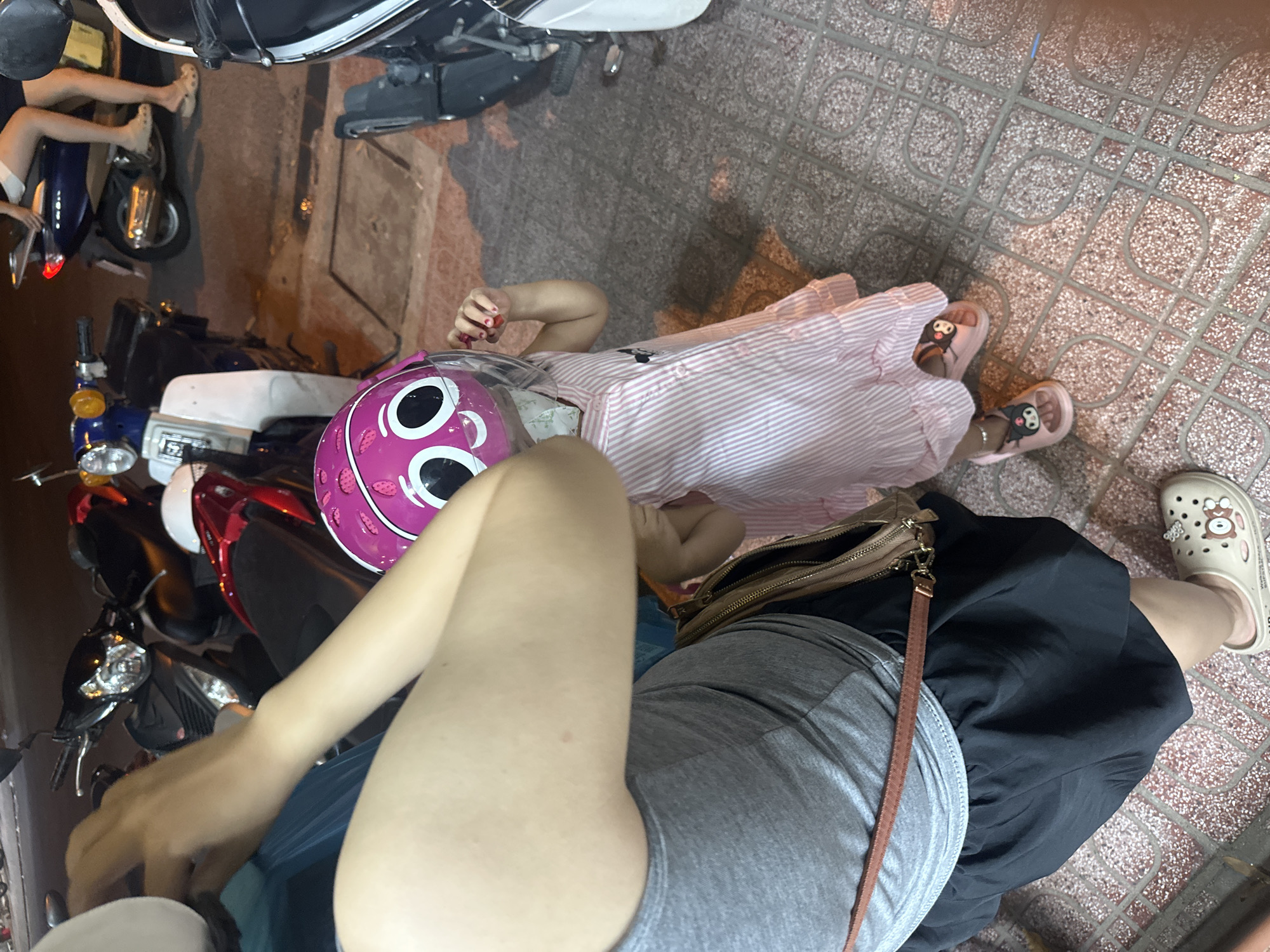 Observed at an elevated, downward angle in Bac Nha Trang Ward, Vietnam, the scene presents an interaction on a tiled street. A child in a pink-and-white striped dress and pink sandals lies near an adult figure, who is dressed in a gray top and black skirt, wearing a tan purse and tan sandals. Both are positioned near a parked red and black motorbike. The child is wearing a pink and white helmet with large round circles on it. A second motorbike is parked near the curb behind the red and black motorbike.
