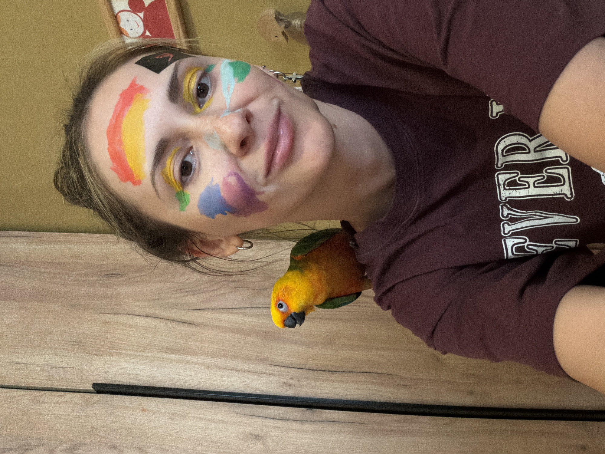 A young woman with paint on her face is holding the camera at an angle, showing a Sun Conure parrot perched on her shoulder. The woman has a rainbow painted above her left eye, yellow paint around both eyes, and blotches of green, blue, and purple paint on her cheek. She has a black square stuck to her forehead. She is wearing a dark maroon-colored sweatshirt. The background includes a wood-patterned wall and an abstract framed image. There is no evidence to support the claim that the image was taken in Khimki, Russia.
