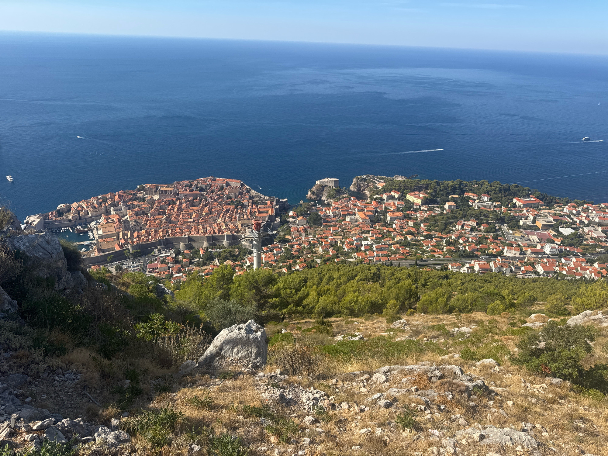 This image captures a panoramic view of the historic walled city of Dubrovnik, Croatia, from a high vantage point on a hillside. The Old Town, characterized by its distinctive red-tiled roofs and stone walls, is prominently situated on a peninsula jutting into the Adriatic Sea. The deep blue sea stretches out towards the horizon, with several small boats visible on its surface. Lush green trees and vegetation cover the slopes in the foreground and surrounding the city, contrasting with the urban landscape. The sky is clear and blue, indicative of a sunny day. The perspective suggests the photographer is positioned on Mount Srđ, a popular viewpoint overlooking the city and its surroundings.