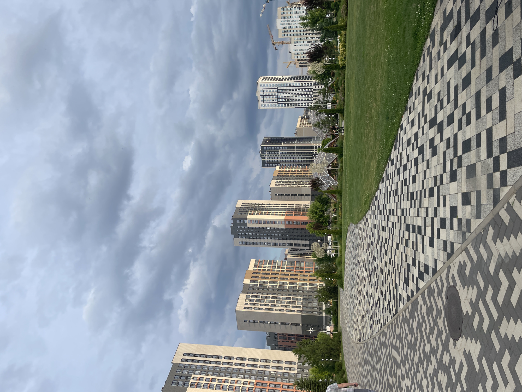 The image shows a modern residential complex in the city of Stavropol, Russia, under a cloudy sky. In the foreground, a winding pathway made of grey and black patterned paving stones leads into a grassy park area. The park features manicured lawns, trees, and some modern architectural elements, including dome-like structures. In the background, several tall, contemporary apartment buildings dominate the skyline, with some construction cranes visible among them, suggesting ongoing development. A person is seen walking on the pathway, and another person is riding a bicycle on the grass in the park. The overall scene depicts a new urban development blending residential buildings with green public spaces.