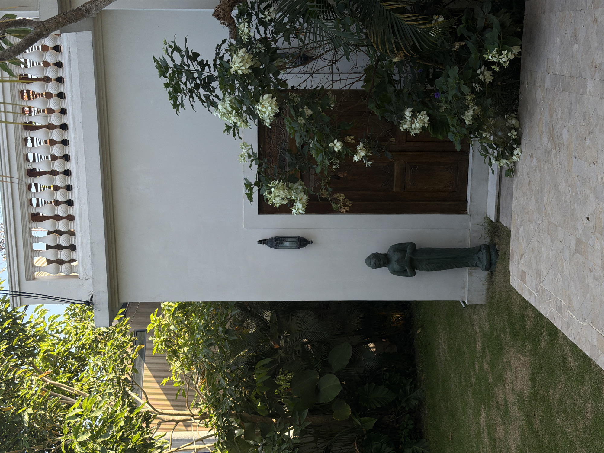 The image depicts a tranquil garden setting outside a building in North Kuta, Indonesia. A lush green lawn stretches across the foreground, leading to a light-colored tiled pathway. To the left of the path, a tall, slender green statue of a person in a meditative pose stands near the corner of a white building. The building features a large, ornate wooden door and a dark, lantern-style light fixture mounted on the wall above and to the left of the door. Adjacent to the door, a flowering plant with numerous white blossoms cascades downwards. Beyond the building, dense foliage and trees create a verdant backdrop, with glimpses of a modern structure with vertical wooden siding visible through the leaves. A white balustrade with decorative pillars adorns the top of the building, suggesting an upper level or balcony. The overall atmosphere is peaceful and serene, characteristic of a well-maintained outdoor space in a tropical environment.