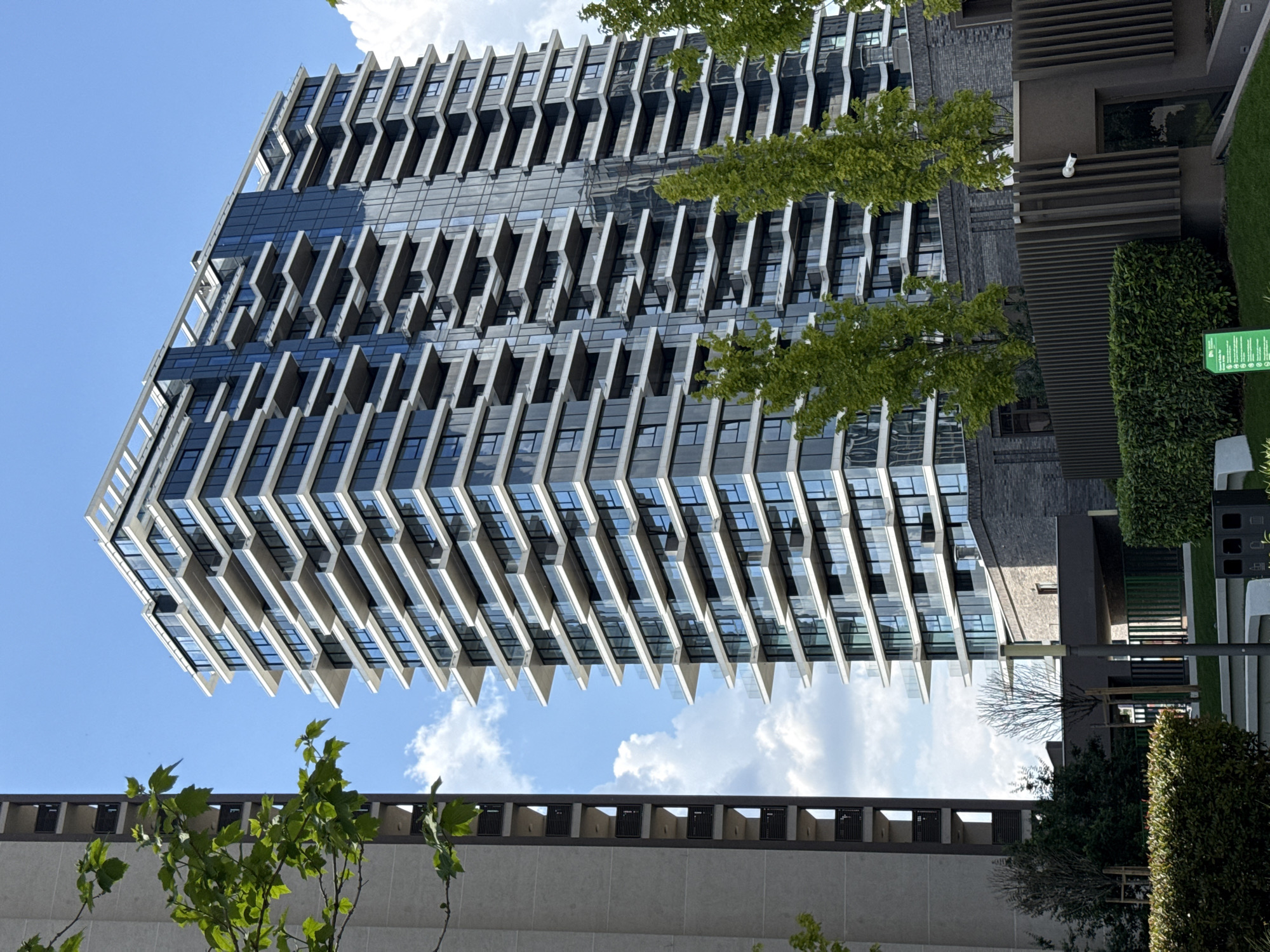 The image shows a modern building located in Belgrade, Serbia. The building has a distinctive design with protruding balconies or sunshades on each floor, creating a zigzag pattern. The structure is primarily made of glass and light-colored concrete or stone. A partial view of the clear blue sky is visible behind the building. Some trees with green leaves are partially visible in the front. Additionally, the top portion of another building is visible in the lower section of the image. A security camera is also attached to the building.