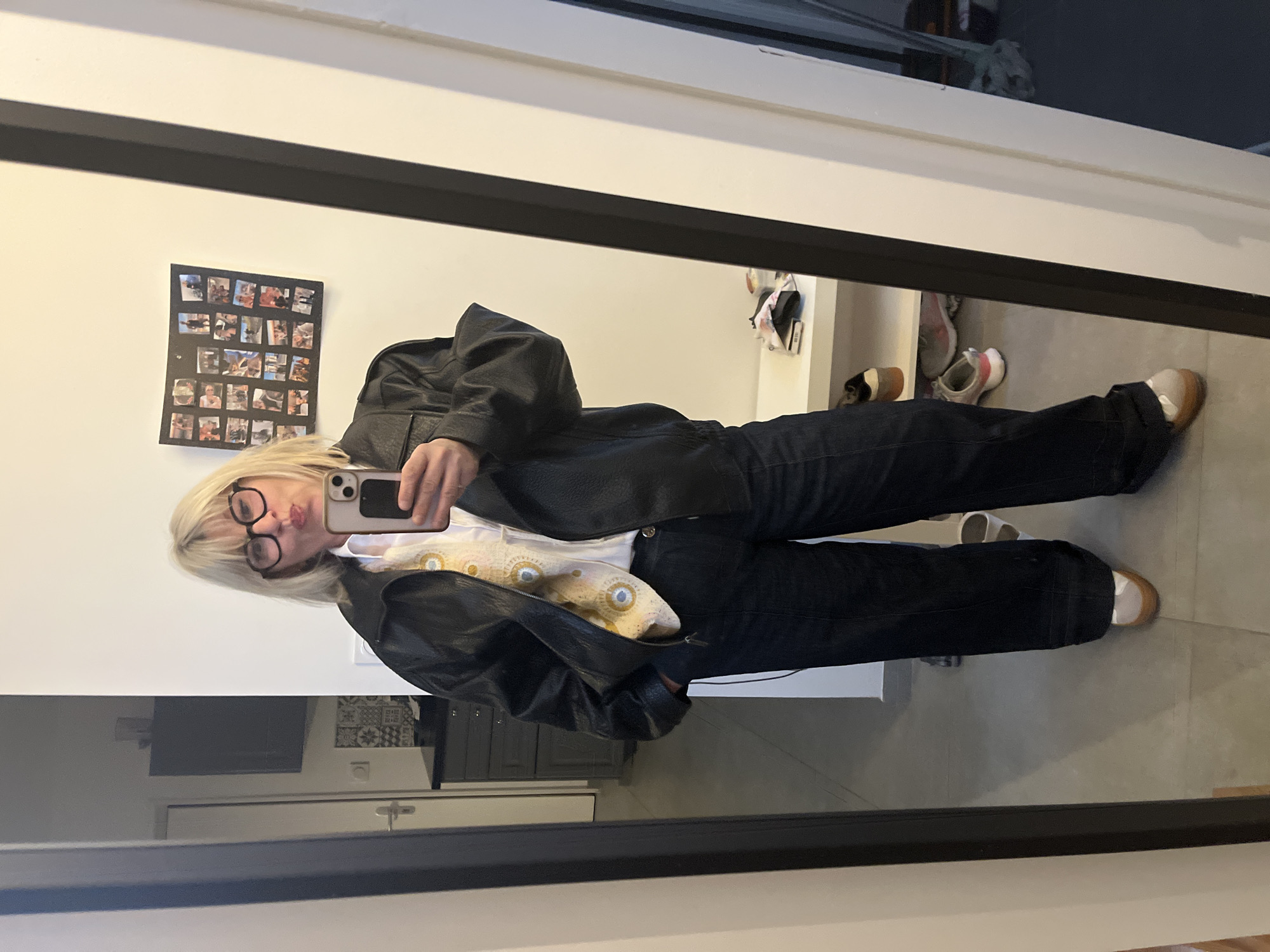 An individual is depicted in a full-length mirror, taking a selfie inside a residence in Chambœuf, France. The subject, an adult with blonde, shoulder-length hair and dark-rimmed eyeglasses, presents with pursed lips while holding a light-colored smartphone with a dark case in their right hand. The individual wears a black leather jacket, open over a white collared shirt with a yellow patterned scarf or tie. Dark blue wide-leg jeans are worn, with the left hand placed in a front pocket. Footwear consists of light-colored, low-top shoes with tan soles.
The mirrored background displays an interior space featuring light grey floor tiling. To the left of the subject, a dark grey cabinet with drawers and a patterned tile backsplash is visible, surmounted by a dark rectangular object, possibly a screen. A white door with a silver handle is partially observed in the deeper background. To the right of the subject, a white wall displays a black framed collage composed of multiple small photographs. Beneath the collage, a white shelf contains several pairs of shoes, predominantly sneakers, and miscellaneous items.