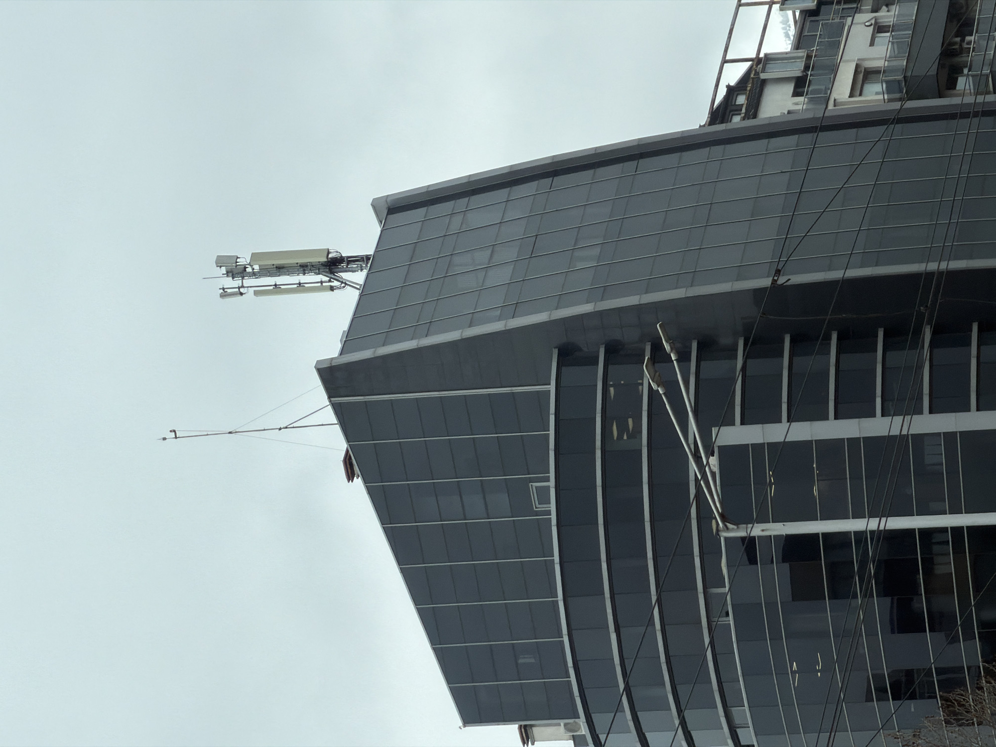 The image captures the upper portion of a modern glass building against a cloudy sky. The building has a distinctive angular design with dark, reflective windows organized in a grid pattern. There are support structures or scaffolding visible around the upper levels, along with communications equipment on the roof. Power lines run in front of the building. The location is the city of Sofia, Bulgaria.