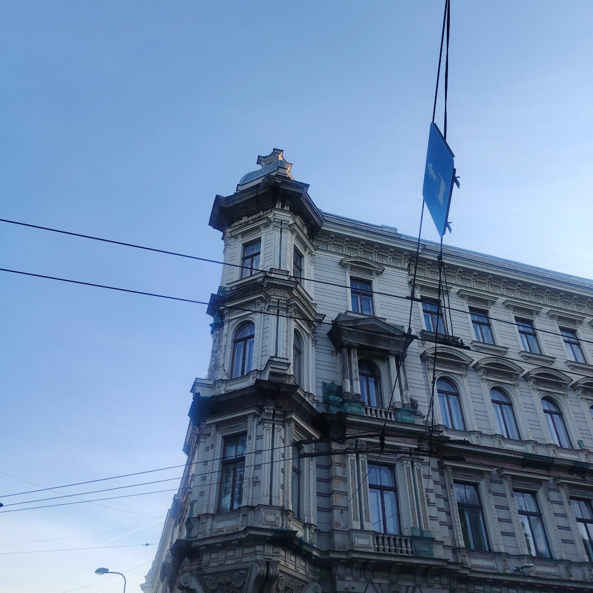 The image shows a corner of a building in Riga, Latvia, under a clear blue sky. The building is an older structure with ornate architectural details, including arched windows, columns, and decorative cornices. A blue sign is hanging from a wire connected to the building. Electrical wires are visible crossing the sky in the background. A street light is visible on the lower left side of the image.