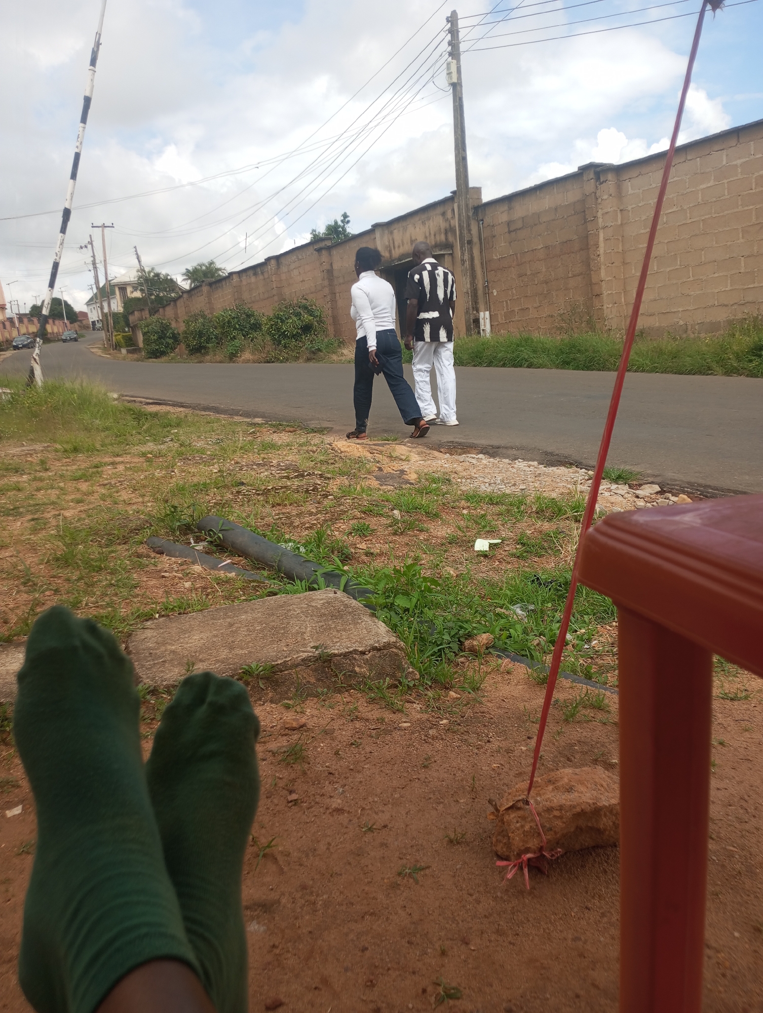 This image depicts a street scene in Jos, Nigeria. In the foreground, a pair of feet wearing green socks is visible, suggesting the perspective of someone relaxing or resting. The background features a paved road with two individuals walking away from the camera. One person is wearing a white top and dark pants, while the other is dressed in a black and white striped shirt and white pants. A tall, striped pole, possibly a traffic barrier, stands to the left, and a brick wall lines the right side of the road. Overhead, power lines crisscross against a cloudy sky. The ground is covered in soil and sparse vegetation, with some concrete elements and dark pipes also present. A red pole, part of a structure out of frame, extends into the scene, with a stone tied to it by pink twine. The overall impression is a casual outdoor setting within the urban environment of Jos.