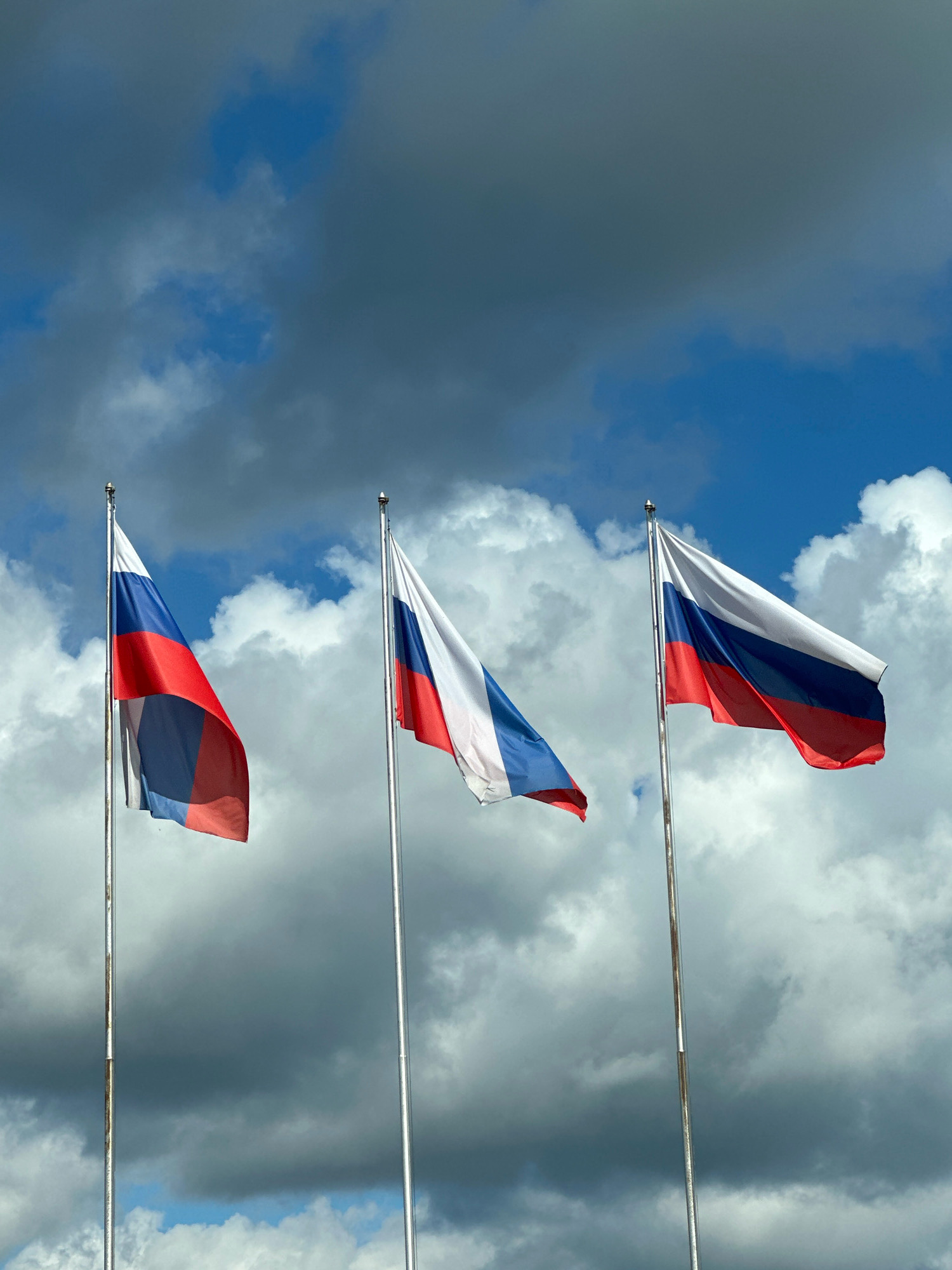 Three Russian flags are displayed on flagpoles against a cloudy sky. The flags are horizontally striped in white, blue, and red. The flag on the left is partially obscured by the flag in the middle, and the flag on the right is fully visible. The flagpoles are silver and appear to be made of metal. The sky is a mixture of blue and gray clouds. The scene is outdoors. The image does not show any people or specific landmarks that would indicate a precise location within Novoivanovskoe, Russia.