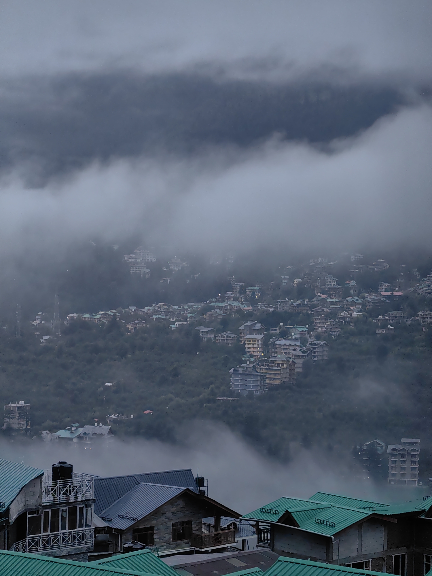 This is a vertical, eye-level, medium shot of the city of Vashisht, India, during a cloudy and foggy day. The scene is dominated by a vast expanse of green hills covered in a thick layer of fog. In the foreground, several buildings with green and dark blue corrugated metal roofs are visible. These buildings appear to be residential or commercial structures, some with balconies and multiple floors. Further into the background, the fog partially obscures a densely populated hillside town. Numerous buildings, varying in size and architectural style, are scattered across the slopes. The fog creates a hazy, ethereal atmosphere, with some of the upper stories of buildings peeking through the mist. The overall impression is one of a remote mountain town enveloped in atmospheric weather conditions. There are no people or animals visible in the image.