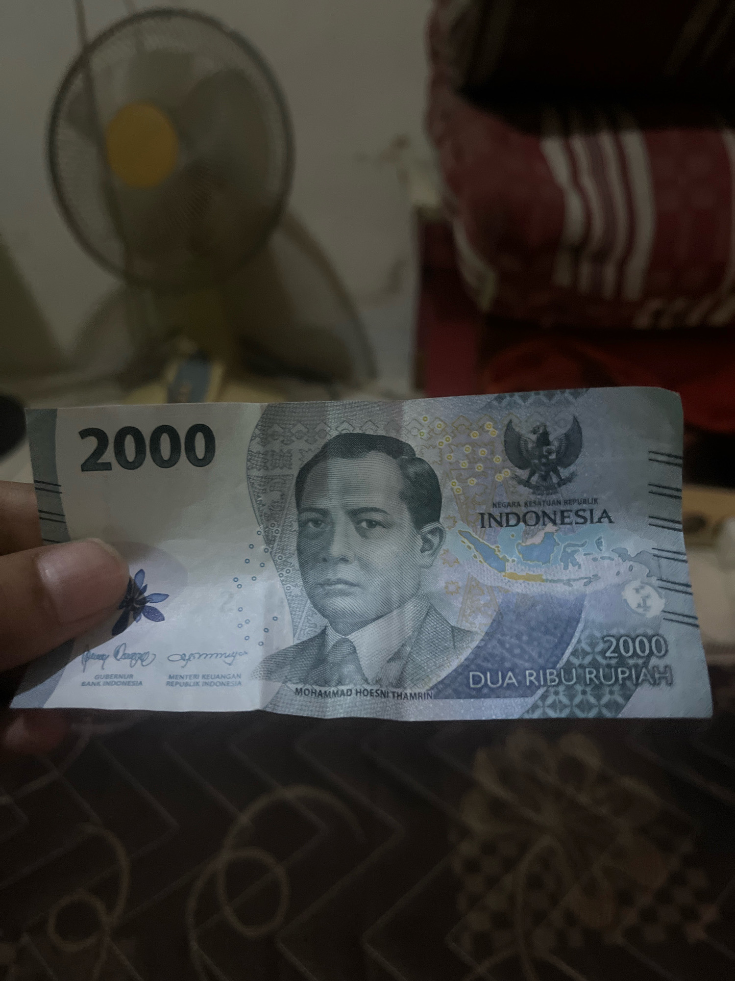A close-up, eye-level, medium shot captures a person's hand holding a Indonesian 2000 Rupiah banknote. The banknote is predominantly gray and white, featuring a portrait of Mohammad Hoesni Thamrin, the national emblem of Indonesia (Garuda Pancasila), and the text "NEGARA KESATUAN REPUBLIK INDONESIA" and "DUA RIBU RUPIAH." The background is blurred, showing a standing fan with a yellow center and a textured surface that appears to be a bedspread with red and white stripes. The scene is indoors, likely in a residential setting within the city of South Jakarta, Indonesia. No specific landmarks or environmental features indicate the precise location within the city.