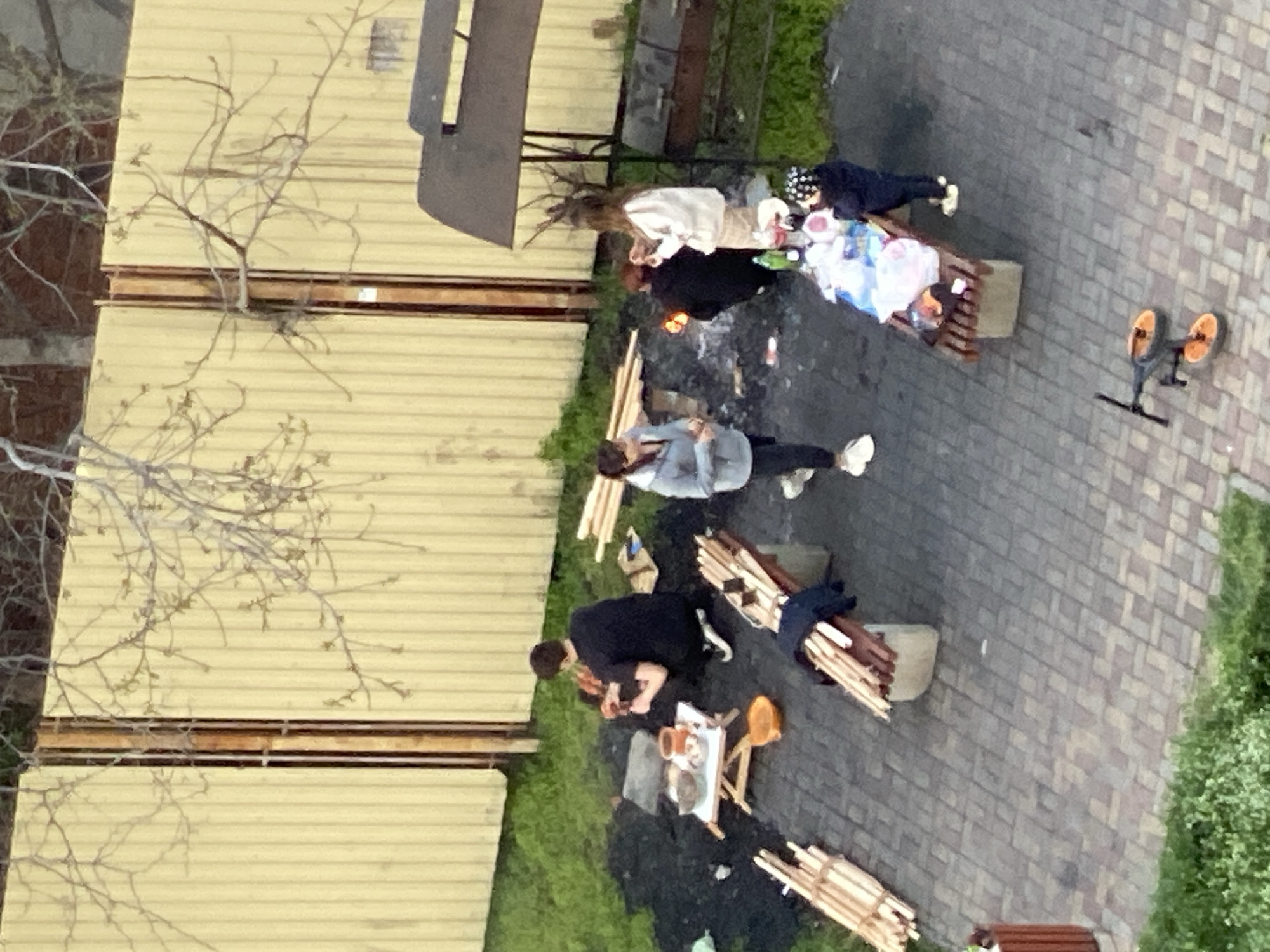 This image, taken from an elevated perspective in Krasnodar, Russia, depicts an outdoor social gathering, likely a barbecue or picnic, in a paved residential or communal area.
Several individuals are present, including adults and potentially a child. In the central foreground, a person wearing a black jacket is crouched near a small fire or grill with glowing embers. Adjacent to this, a small, light-colored table is set with cups and dishes, suggesting food and drinks are being served. Bundles of thin wooden sticks, possibly kindling or fuel, are stacked on the ground nearby and on a low wooden stand.
Two wooden benches with metal frames are visible. One bench, positioned to the right, holds various items including plastic bags and bottles. A person in a dark blue jacket and a black head covering with white dots stands near this bench, alongside what appears to be a child or additional items. Another individual with long hair, wearing a light-colored top, stands near the second bench. Further into the scene, a person in a grey-blue top and dark pants is also present.
The ground is paved with grey interlocking bricks, and a strip of green grass is visible along one side. A light-colored fence or wall, composed of horizontal panels, runs along the left side of the scene, with bare tree branches silhouetted against it, suggesting late winter or early spring. An orange and black kick-scooter or bicycle with two large orange wheels is parked on the paved area towards the right. The lighting appears somewhat dim, indicating late afternoon or early evening. No visible text is discernible. The scene conveys a relaxed outdoor social event.