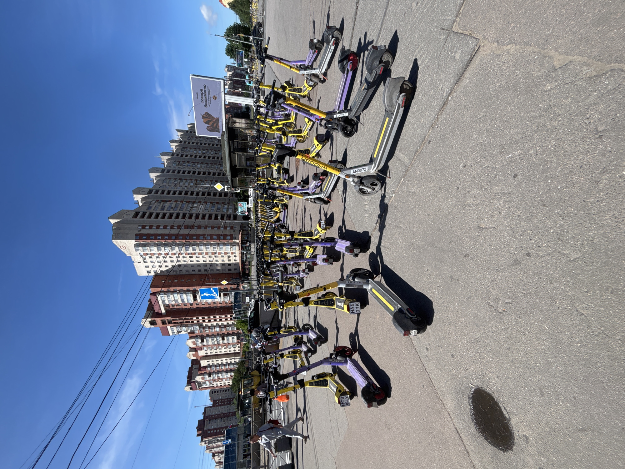 An outdoor, eye-level, medium shot captures a large group of parked electric scooters on an asphalt surface. The scooters, predominantly yellow and purple with some gray accents, are lined up facing various directions. Some scooters have license plates visible, displaying alphanumeric characters such as "HC8 94X," "HA6 01K," and "ANQ072." In the background, a woman in gray athletic wear walks from left to right, and a construction vehicle is visible. The scene is framed by multi-story apartment buildings with red and white facades, and a clear blue sky overhead. A large billboard with Russian text and an image of a bee is also present in the background. The environment appears to be a city street or public space.