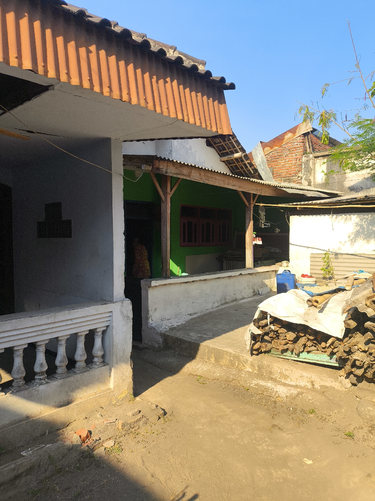 The image depicts an outdoor scene in what appears to be a residential area in Labruk Kidul, Indonesia. The foreground features a dirt and concrete ground surface with scattered debris, including broken bricks and patches of dry grass. To the right, a large pile of chopped firewood is covered with a white tarp. Behind the firewood, a green metal structure is visible.
In the mid-ground, a pathway made of concrete and steps leads towards a building with a green wall and a wooden-framed roof extension. A person, partially obscured by shadow, is visible standing in the doorway of the green building, wearing a patterned garment. A large blue plastic container is situated on the ground near the steps. Adjacent to this building is another structure with a white wall and a corrugated metal roof.
The background reveals more of the residential architecture, including terracotta tiled roofs and exposed brickwork on one building. A portion of a tree with green leaves and thin branches extends into the upper right corner of the frame. The sky is clear and bright blue. The lighting suggests it is daytime, with strong shadows cast by the buildings and objects. The overall impression is of a somewhat unkempt but lived-in outdoor space.