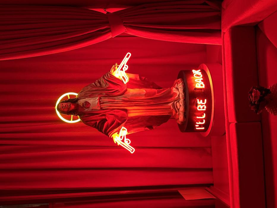 A Jesus statue is surrounded by a red background. The statue features a golden halo and is holding neon pistols. The statue sits atop a rotating base with a brain depicted on the sides. The words "I'LL BE BACK" are illuminated in neon lettering. A red curtain and what appears to be the side of a sofa are visible. The lighting is red, casting a dramatic and bold ambiance.