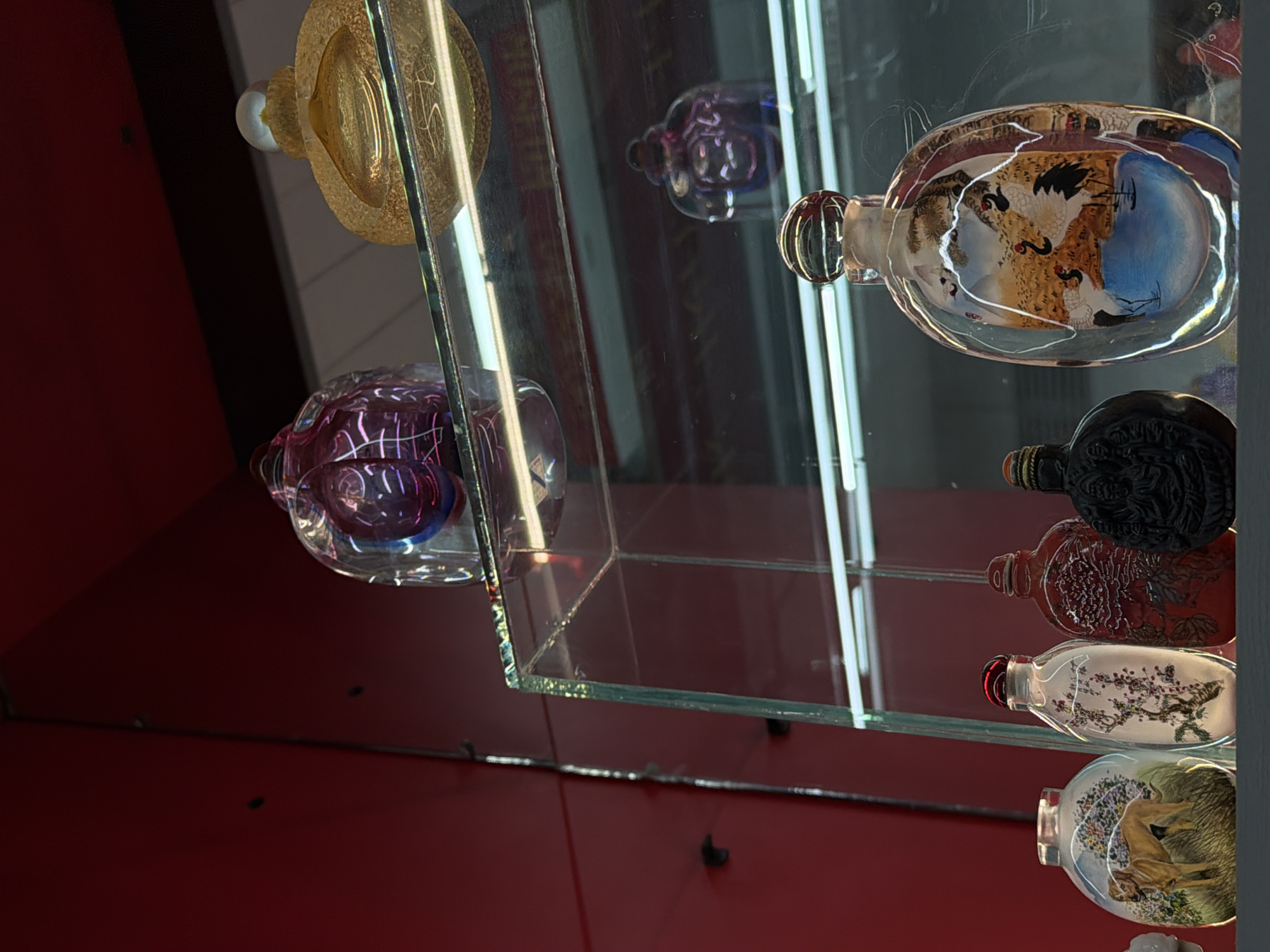 The image displays a collection of decorative bottles, primarily of the snuff bottle type, arranged within a multi-tiered glass display cabinet. The background of the cabinet is a uniform, dark red color. Artificial light sources are visible as horizontal reflections on the glass shelves and bottles.
On the uppermost visible glass shelf, a clear, faceted, spherical object with internal purple and blue swirling patterns is positioned on the left. To its right, a textured, golden-yellow, flattened spherical bottle with a clear, spherical stopper is present.
The lower, prominent glass shelf features five distinct bottles, from left to right:
1. A clear, flattened oval bottle containing an internal painting of a light brown dog standing in a green, grassy landscape with flowering bushes.
2. A clear, slender oval bottle with an internal painting of a pink cherry blossom branch on a white background, topped with a translucent red stopper.
3. A dark red, opaque bottle with a textured surface featuring raised, intricate patterns resembling foliage or blossoms, with a matching dark red stopper.
4. A dark-colored, opaque bottle with a detailed carved relief of a seated figure on its front, secured with a dark stopper.
5. A larger, clear, flattened oval bottle showcasing an internal painting of multiple white and black cranes near a blue body of water, against a landscape of brown hills and sparse trees, topped with a clear, spherical stopper.
Reflections of the surrounding environment and internal lighting are visible on the glass surfaces of the cabinet and the bottles. No specific identifying features are present to determine the precise location within the city of Yerevan, Armenia.