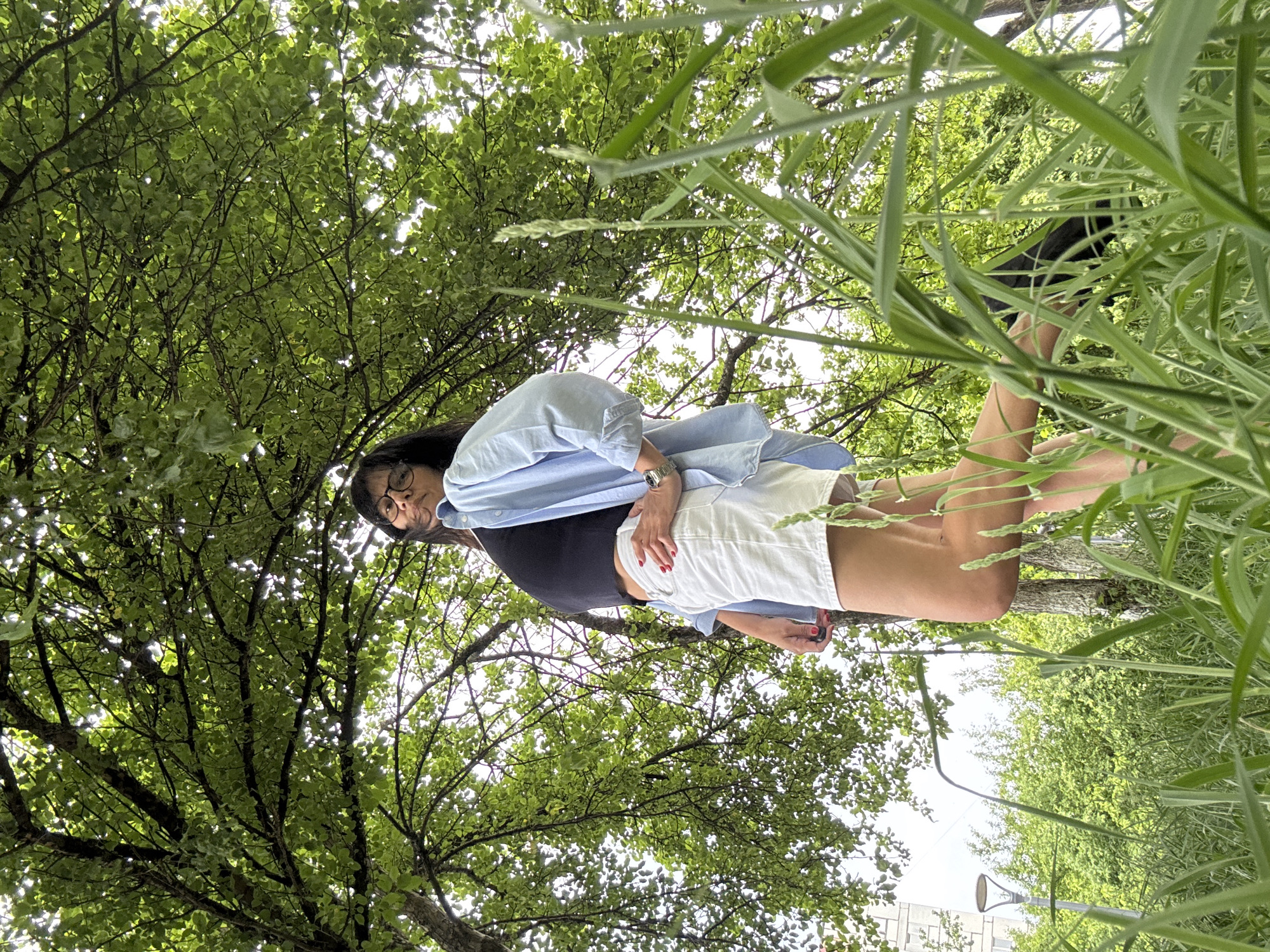 A person with glasses, a black tank top, a light blue button-down shirt, and white shorts is lying on the ground, looking upward. The shot is taken from a low angle, looking up through tall grass and tree branches filled with green leaves. In the background, a streetlight and a building can be seen. The location appears to be a park or grassy area within a city.