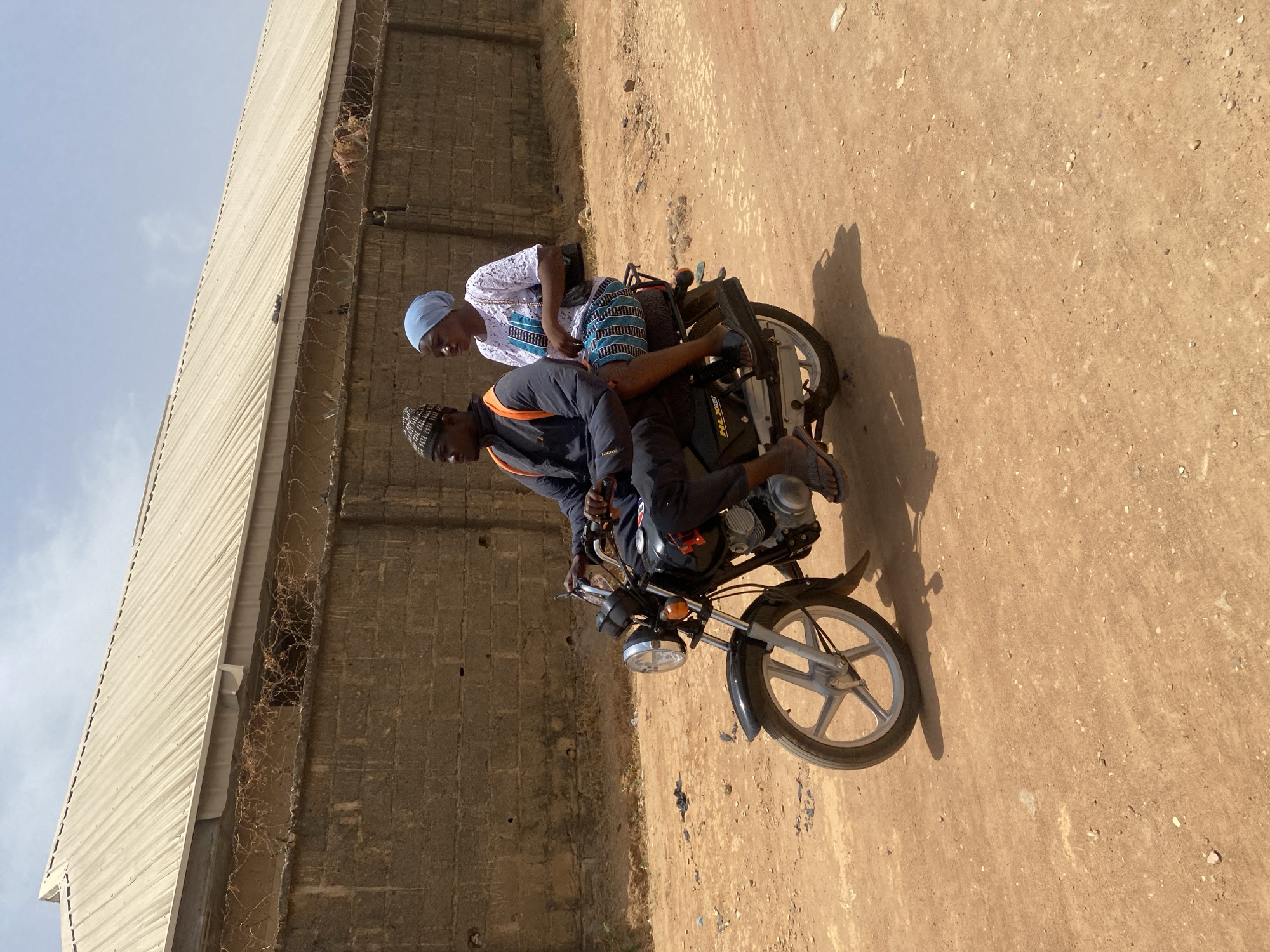 Two individuals are on a black motorcycle on a dirt road. The person in front is wearing a dark jacket with an orange stripe, dark pants, sandals, and a patterned kufi cap. The person in the back is wearing a white shirt, blue and white patterned pants, sandals, and a light blue kufi cap. The background includes a tall, brick wall topped with barbed wire. The ground is a mix of dirt and gravel. The location appears to be Rido, Nigeria.