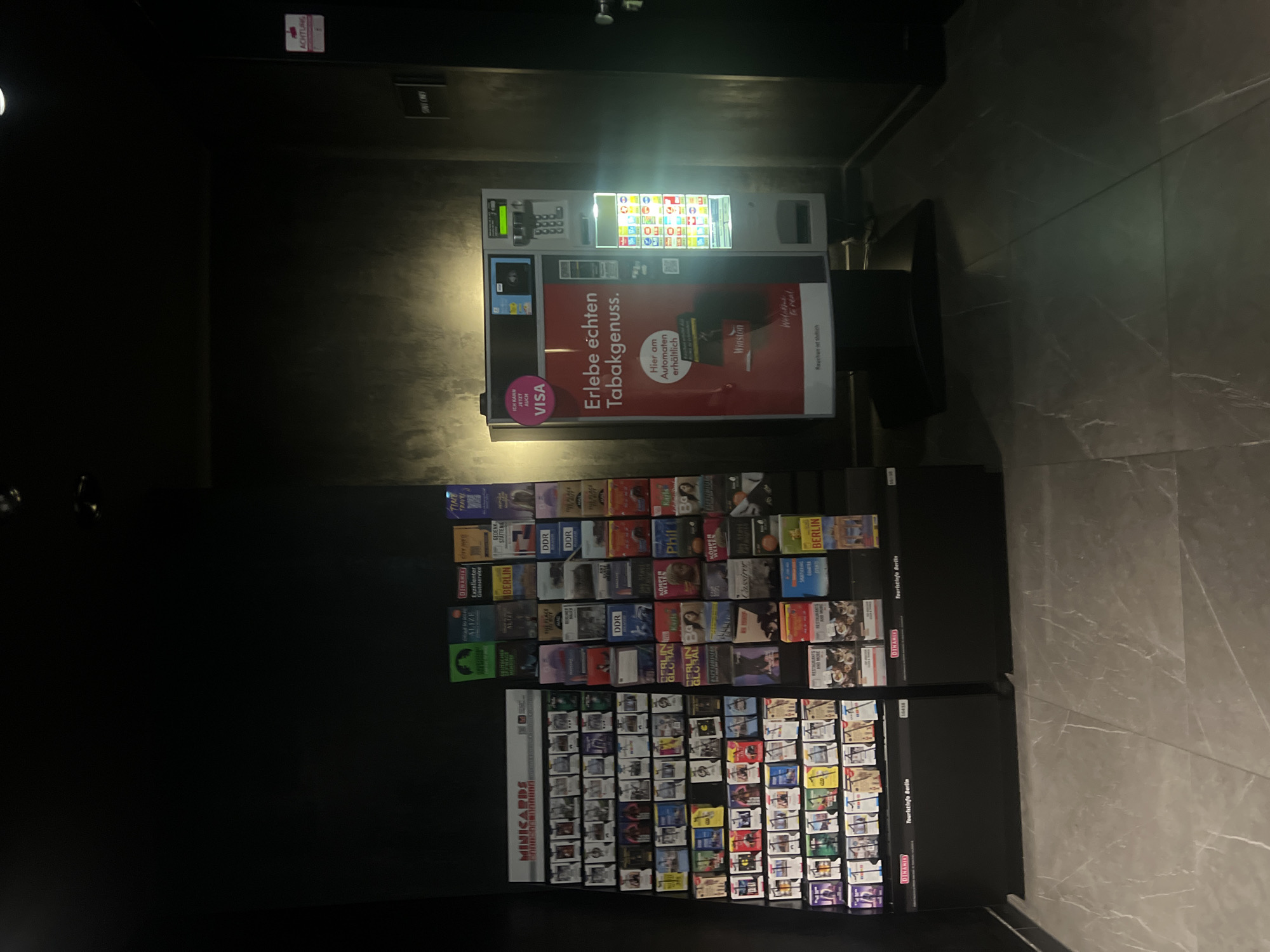 This is a medium shot, eye-level, indoor photograph of a public space, likely a tourist information area or a transit hub in Berlin, Germany. The scene is dimly lit, with a dark, textured wall forming the background. To the left, a display rack is filled with numerous small, colorful brochures and minicards, organized into many rows and columns. These appear to be promotional materials for various attractions, services, or locations, with some labels visible including "MINICARDS", "DDR", "BERLIN GLOBAL", and "PHIL". Below the brochure rack are two black cabinets, one labeled "DINAMIES Touristinfo Berlin."
To the right of the brochure display stands a large, modern vending machine. The machine has a prominent red front with text in German, including "Erlebe echten Tabakgenuss." (Experience real tobacco enjoyment). There's a section for a Visa payment option, and the machine displays various product images and a keypad for selection and payment. The overall environment suggests a commercial or informational setting designed for tourists.