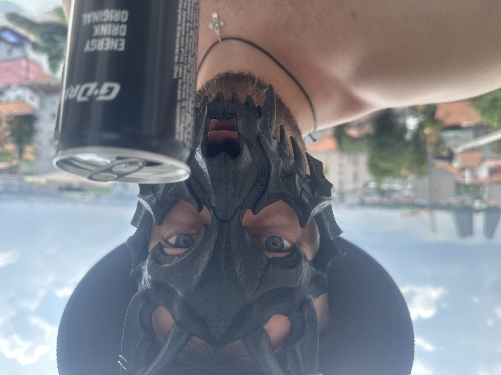 The image shows a close-up, upside-down selfie of a person wearing a black, spiky mask that covers their face, with only their eyes and mouth visible. The mask has a somewhat demonic or monstrous appearance. The person has blue eyes and a light complexion, with short brown hair and facial hair visible around the mouth area. They are wearing a black necklace with a small silver cross pendant. To the left of the mask, a black energy drink can is partially visible, with text that reads "ENERGY DRINK ORIGINAL" and "G-DRIVE" in white lettering, oriented vertically.
The background appears to be an outdoor scene, with blurred buildings and greenery suggesting an urban or suburban setting, possibly with a body of water in the distance. The sky is visible with white clouds, indicating it is daytime. The overall mood is mysterious and slightly unsettling due to the mask. The location is identified as Zvezdara Urban Municipality, Serbia.