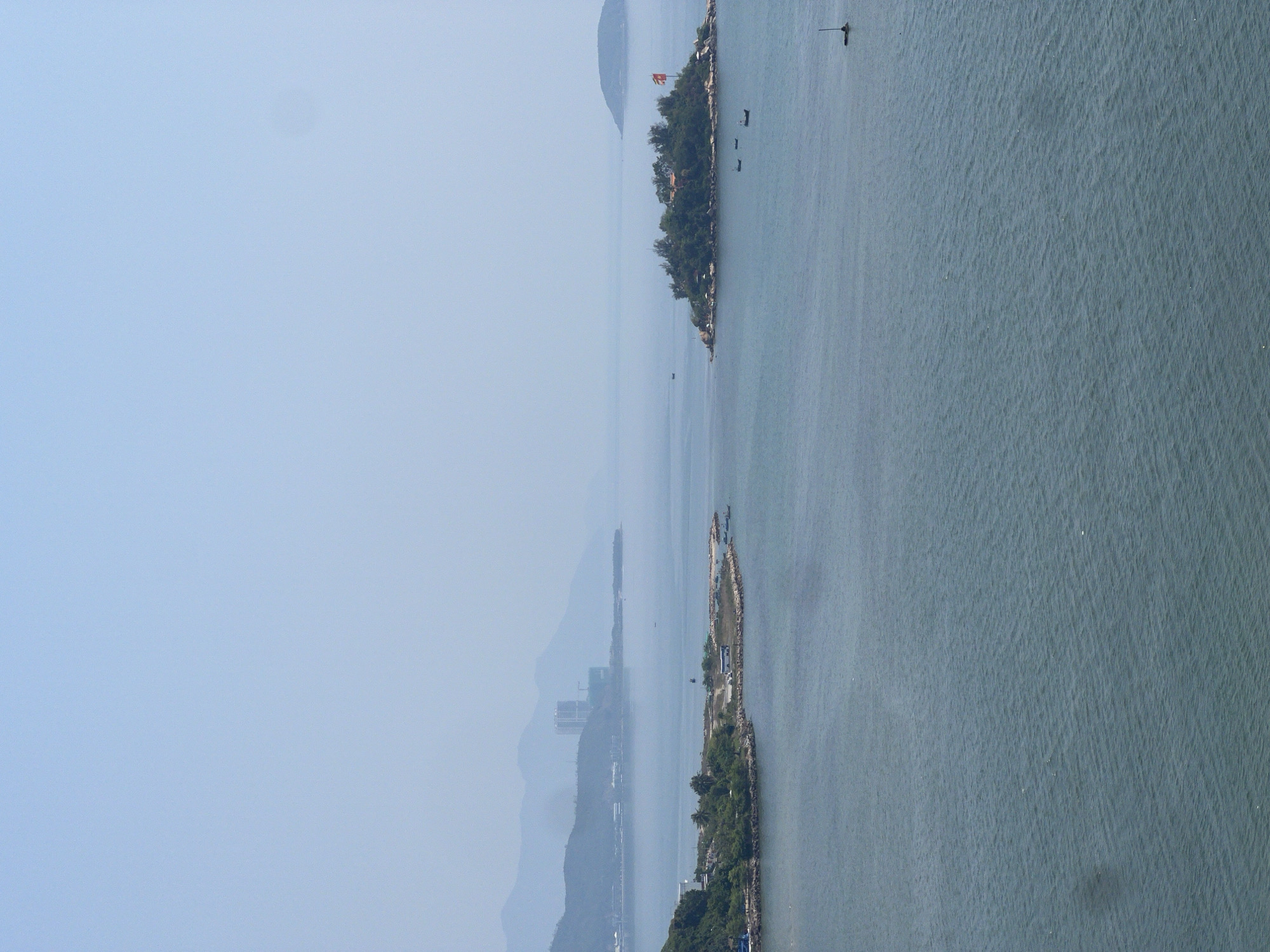 This aerial shot depicts a seascape, possibly taken near Nha Trang, Vietnam, featuring two prominent islands situated within a body of water. The sky above is clear and blue. The water is calm. The first island is lush with vegetation and features a small structure flying a flag. The second island shows signs of infrastructure, including a road or pier-like extension, suggesting human activity. The mainland, possibly a part of Nha Trang, is visible in the distance, characterized by buildings and mountainous terrain shrouded in a light haze.