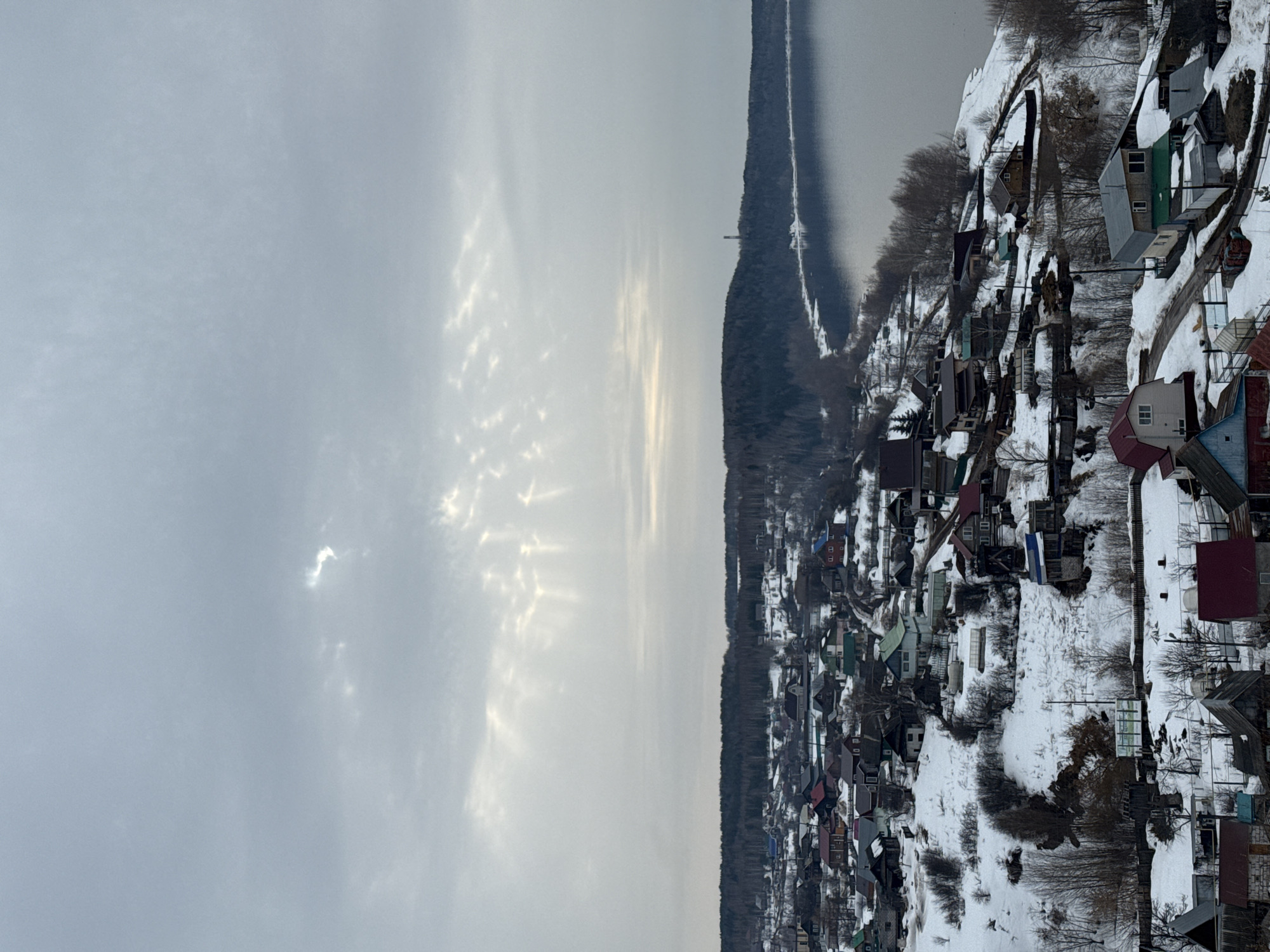 The image presents an elevated panoramic view of a snow-covered residential settlement, identified as Ольховка, Russia, situated on a hillside descending towards a large body of water. The sky is uniformly overcast and gray, with several distinct crepuscular sun rays visible descending through cloud breaks in the upper central portion of the frame.
The foreground and midground are dominated by numerous single-story and multi-story houses of varied construction, featuring roofs in colors such as red, green, blue, brown, and gray. These structures are scattered across a terrain heavily covered in snow, interspersed with areas of exposed brown soil and dormant deciduous trees and shrubs. Unpaved paths or roads are visible winding through the snow-covered landscape between the dwellings.
To the right, a wide expanse of calm water, reflecting the dull sky, extends into the background. The opposite shore of this water body is densely forested with dark, bare trees, and a slender, dark tower or mast is visible rising above the distant tree line on the far bank in the right midground. No human figures or discernible activities are present in the scene.