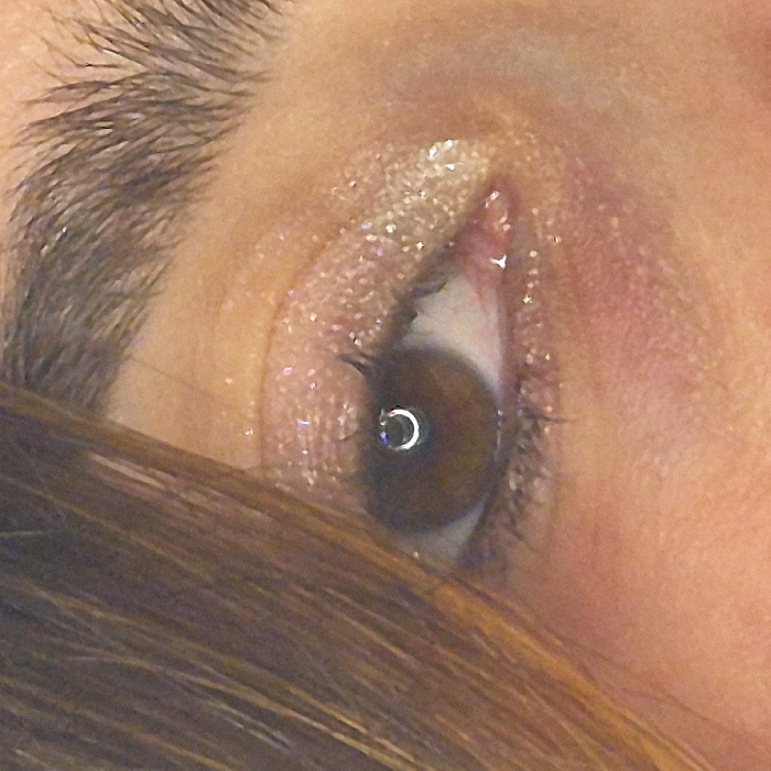 A close-up image reveals an eye with brown iris, surrounded by subtle, shimmery eyeshadow. The brow is thick and well-defined. Sparse eyelashes frame the eye. A strand of brown hair partially obscures the lower portion of the image. The skin around the eye appears fair, with a slightly pinkish hue. The lighting suggests a direct source, reflected as a crescent on the iris.