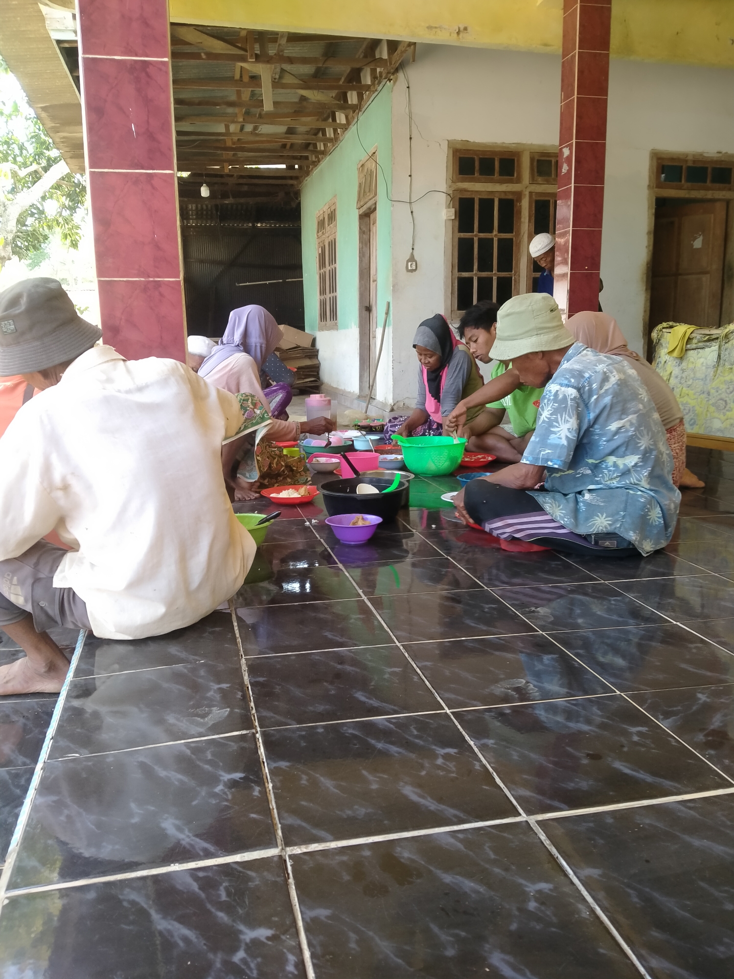 In Blaban, Indonesia, a group of people are gathered on a tiled floor, eating a meal outdoors. The setting appears to be a covered patio or verandah of a building with an unfinished wooden roof structure. To the left, a person wearing a hat and a light-colored shirt is seen from behind, sitting on the floor. In the center, several individuals, some wearing headscarves, are engaged in eating. They are surrounded by various bowls and plates containing food, and a large green colander is also present. The floor is tiled with dark, glossy tiles, reflecting the overhead light. The background shows the exterior wall of a building with windows and doors. The overall scene suggests a communal meal shared in a casual outdoor setting.