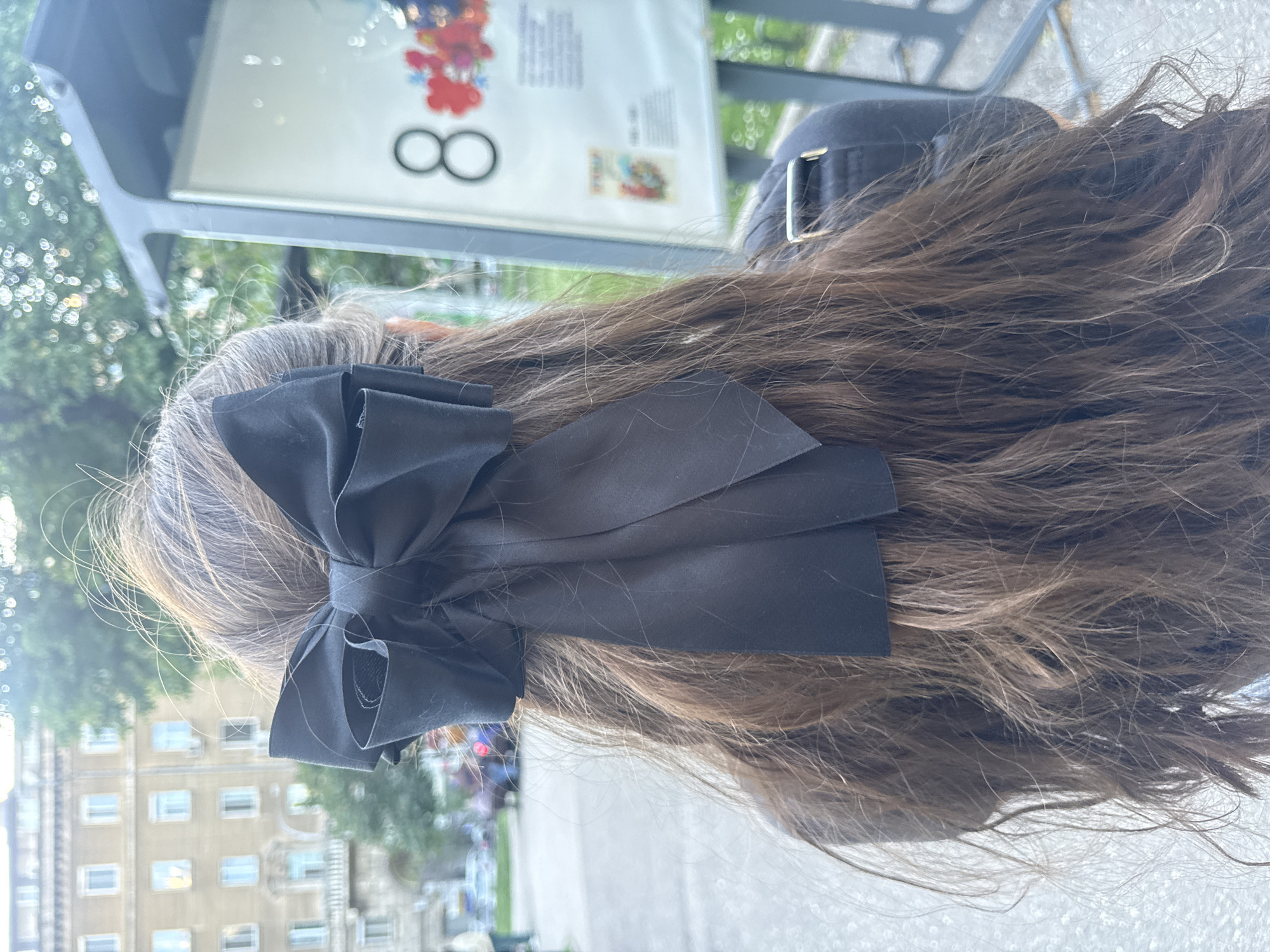 The image depicts the back of a person's head, featuring a large black bow attached to their brown and gray hair. The person is wearing a black cap. In the background, partially visible, is a bus stop sign with an advertisement featuring a stylized "8" and possibly other images. Through the bus stop, buildings and trees are visible, suggesting an urban environment. Based on the architectural style of the buildings, the image appears to be taken in Sofia, Bulgaria.