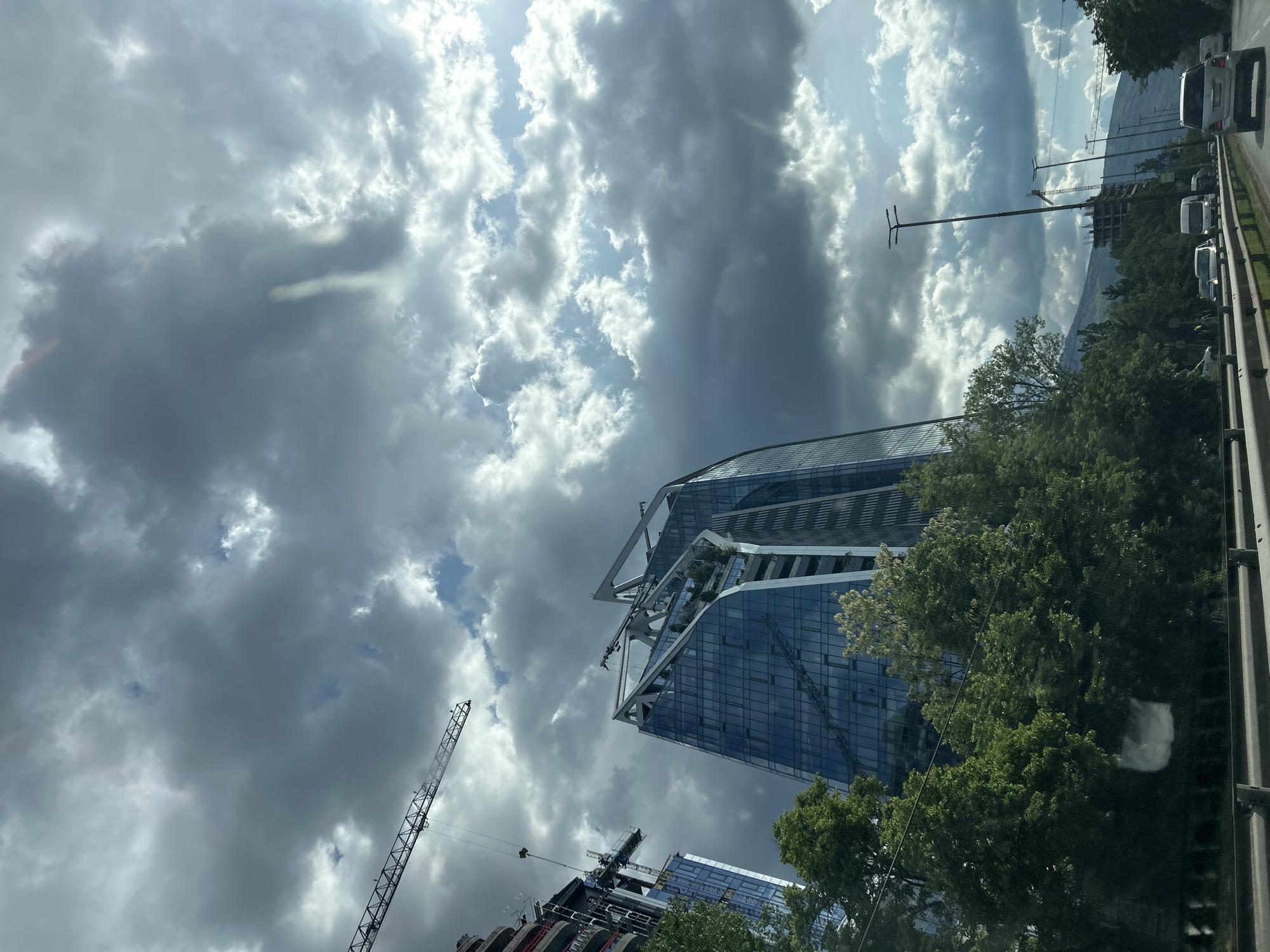 The image shows a view upward towards the sky, buildings, and construction cranes in Sofia, Bulgaria. The sky is filled with a mix of dark, ominous clouds and patches of bright sunlight. Two high-rise buildings with modern architectural designs are visible. The building to the upper right is a tiered structure with reflective glass panels and visible metal support beams. Adjacent to this building are green trees and a road with cars traveling on it. A construction crane is seen in the lower-left corner, along with another building under construction.