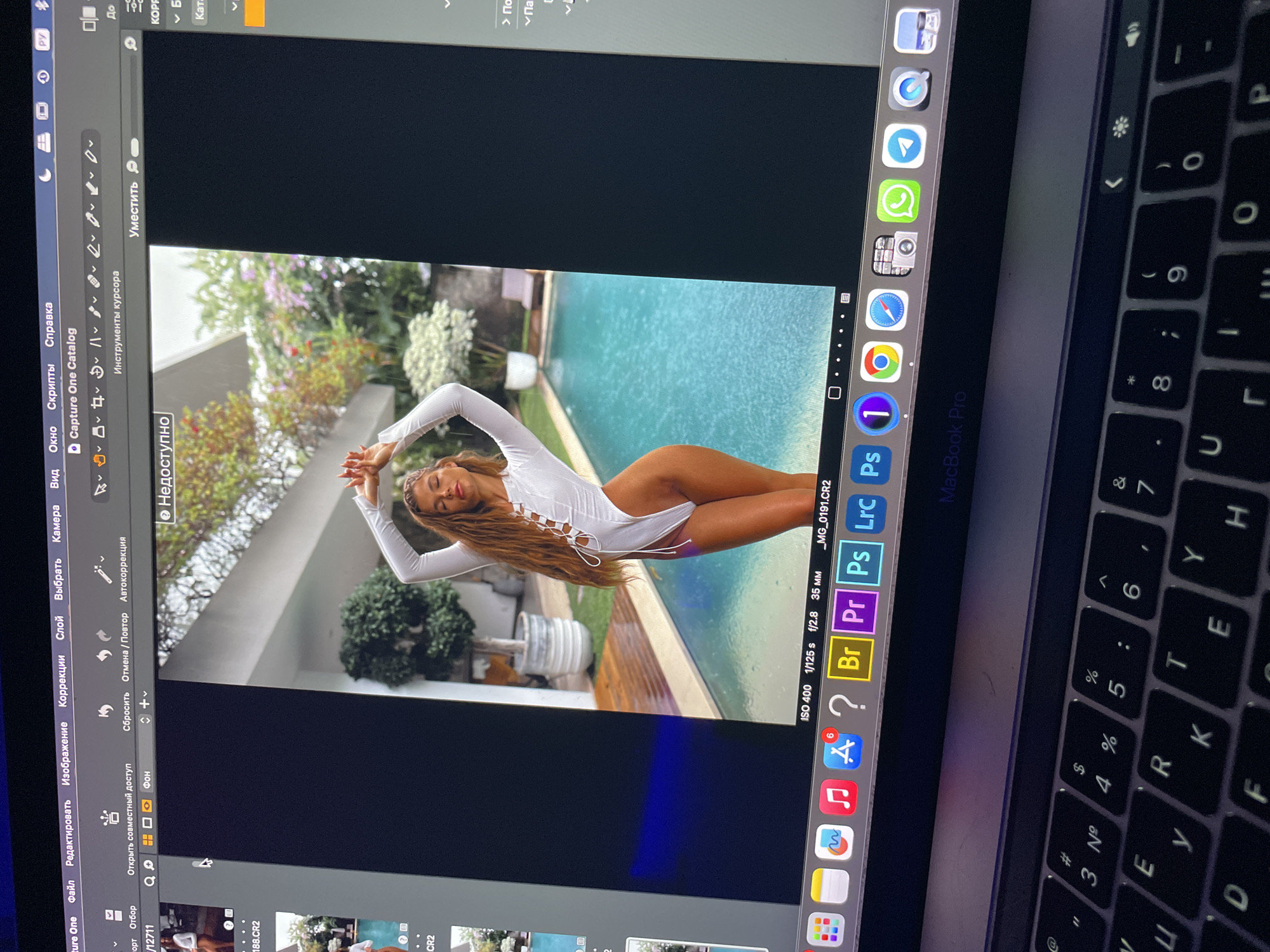 The image is a photo of a MacBook Pro screen displaying a photo editing software, Capture One, opened in Russian. A photograph of a woman in a white swimsuit is being edited. The woman is positioned near a swimming pool and some greenery. The MacBook is also displaying the Dock with icons for applications like Safari, WhatsApp, PhotoShop, and other editing software. Part of the keyboard is visible in the shot.
