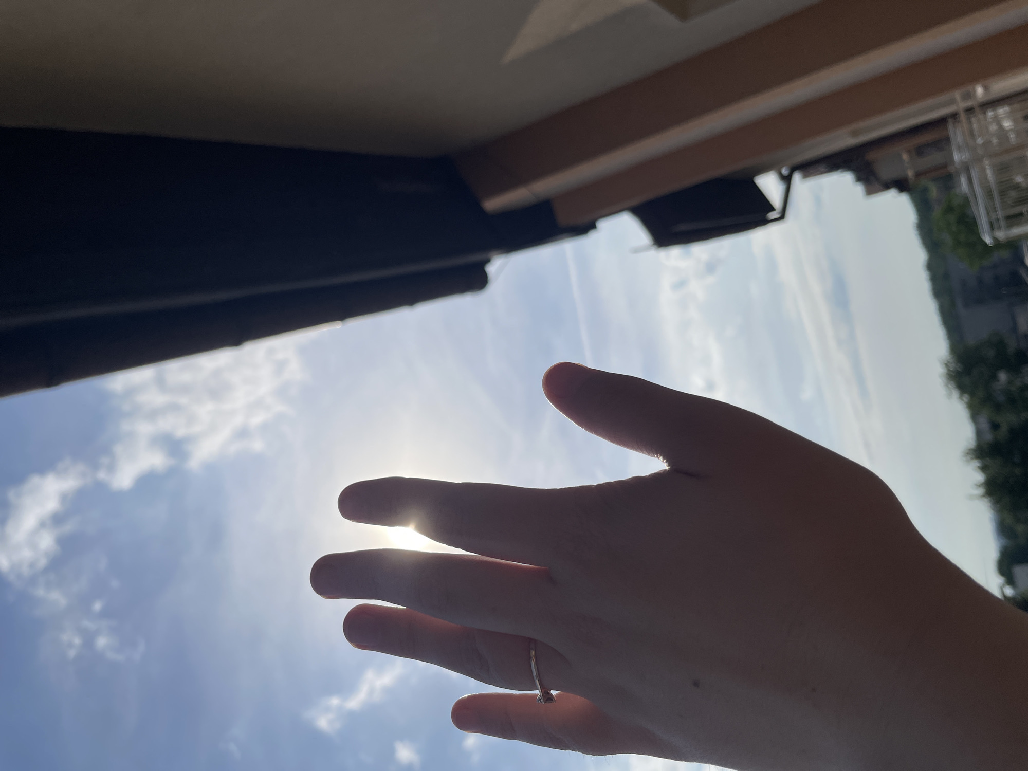 A person's left hand is held up against a bright, sunny sky with scattered clouds. The sun is directly behind the fingers, creating a lens flare effect and silhouetting the hand. A thin, silver ring is visible on the ring finger. To the right of the hand, a dark overhang and a beige building are partially visible. In the background, out of focus, are trees and buildings, suggesting an urban or suburban setting. The location is identified as Unknown, Unknown.