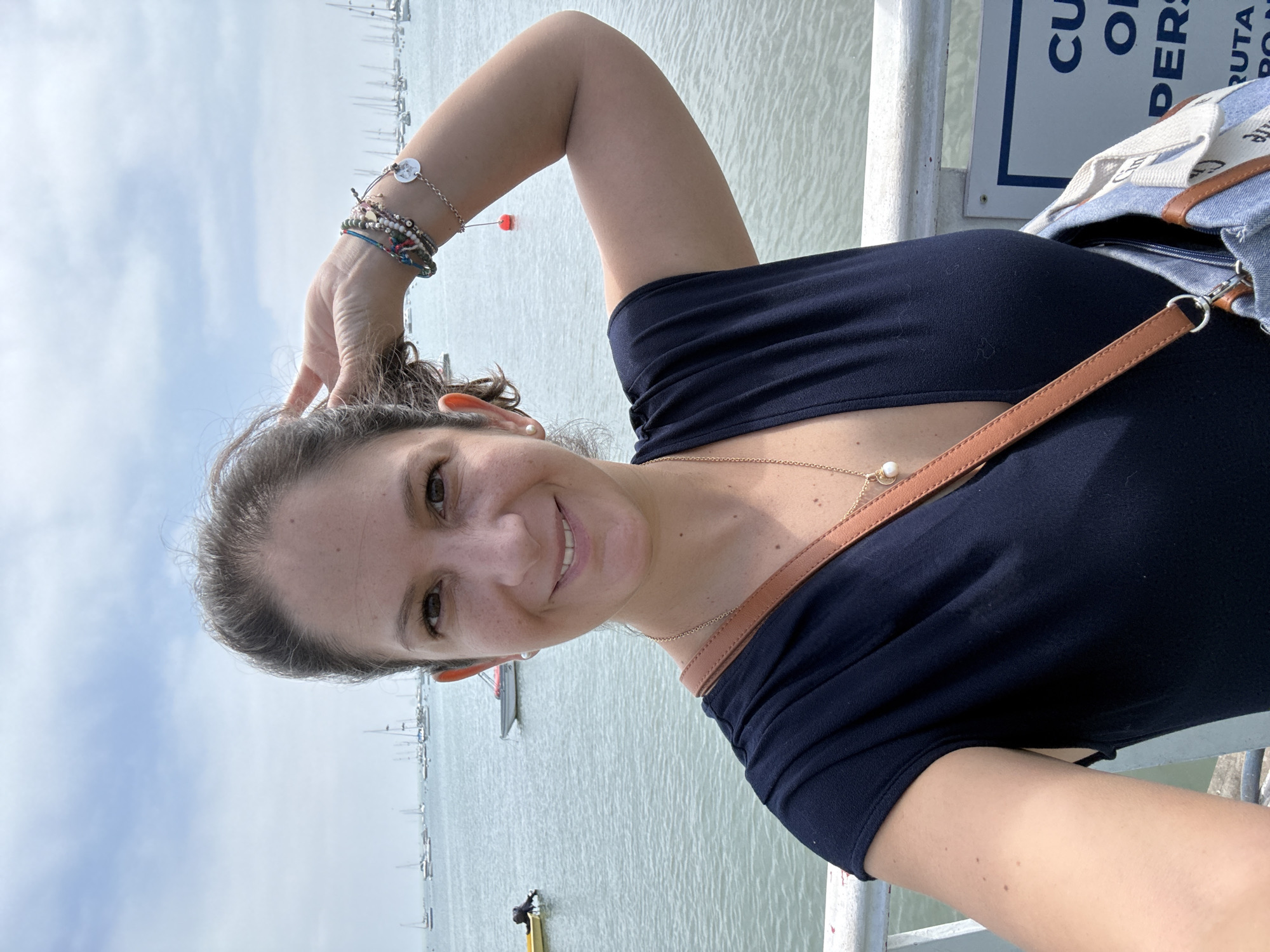 A medium shot of a woman is visible against a backdrop of water and boats. The woman is looking at the camera and is smiling. She has medium to dark hair and skin with dark eyes. She is wearing a dark shirt and jewelry on her wrist. The water behind her shows several boats in the distance. The background has a sky that is mostly cloudy. The photo was likely taken on a pier or dock. The location of the photograph is near La Punta, Peru, based on the presence of sailboats and a pier structure.