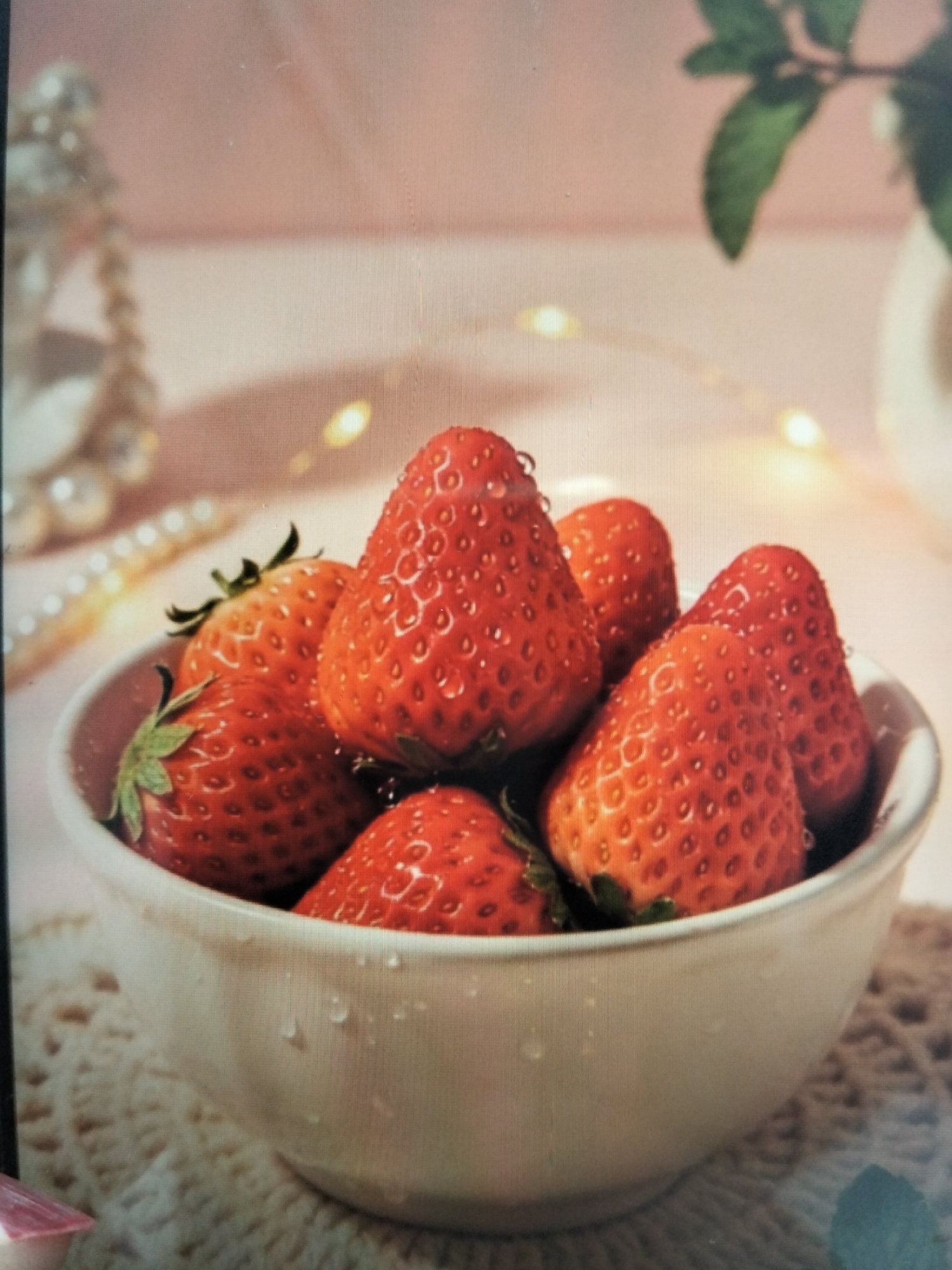 A close-up, slightly out-of-focus photograph depicts a white ceramic bowl filled with ripe strawberries. The strawberries are a vibrant red, with visible seeds and green stems. Some strawberries have water droplets on them, suggesting they are freshly washed. The bowl is placed on a textured, light-colored surface, possibly a doily or placemat, which has a crocheted pattern. In the background, the scene is softly lit with blurred fairy lights, creating a warm and inviting atmosphere. To the left, a decorative strand of pearls and a decorative object, possibly a tiered stand with pearls, are also visible, adding to the elegant presentation. A green leaf, possibly from a plant, is partially visible in the upper right corner. The image does not contain any identifiable people or a specific location in the city of Payagaboh, Indonesia.