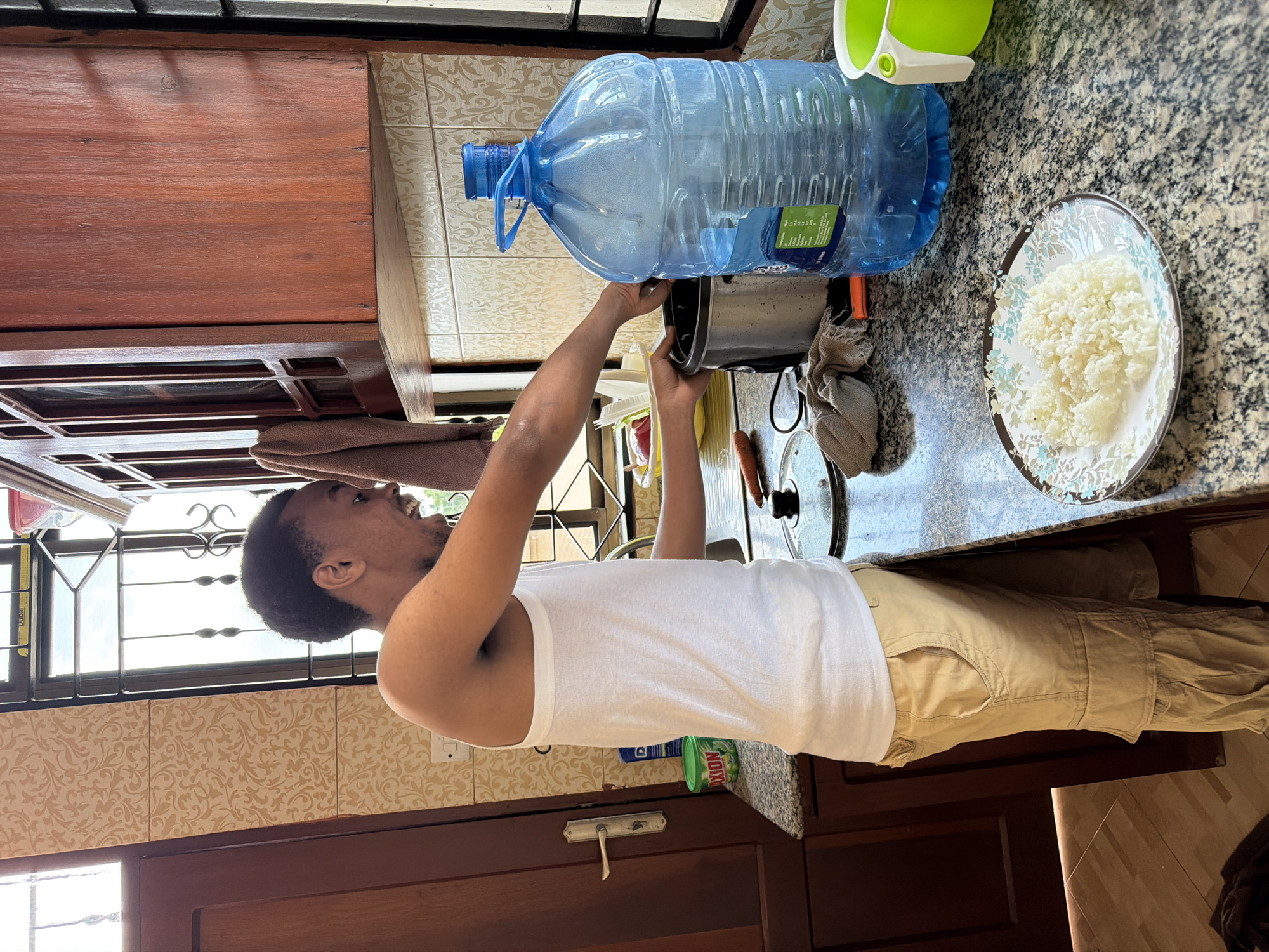 The image depicts an indoor scene, likely a kitchen, in Dar es Salaam, Tanzania. A young man wearing a white tank top and tan cargo pants is standing at a granite countertop. He is in the process of preparing food, with his hands inside a rice cooker. On the counter, there is a large blue water jug, a plate of cooked white rice, a green plastic container, and various kitchen utensils. The background shows wooden cabinets and patterned tiles, characteristic of domestic interiors in the region. Sunlight streams in from a window, illuminating the scene.