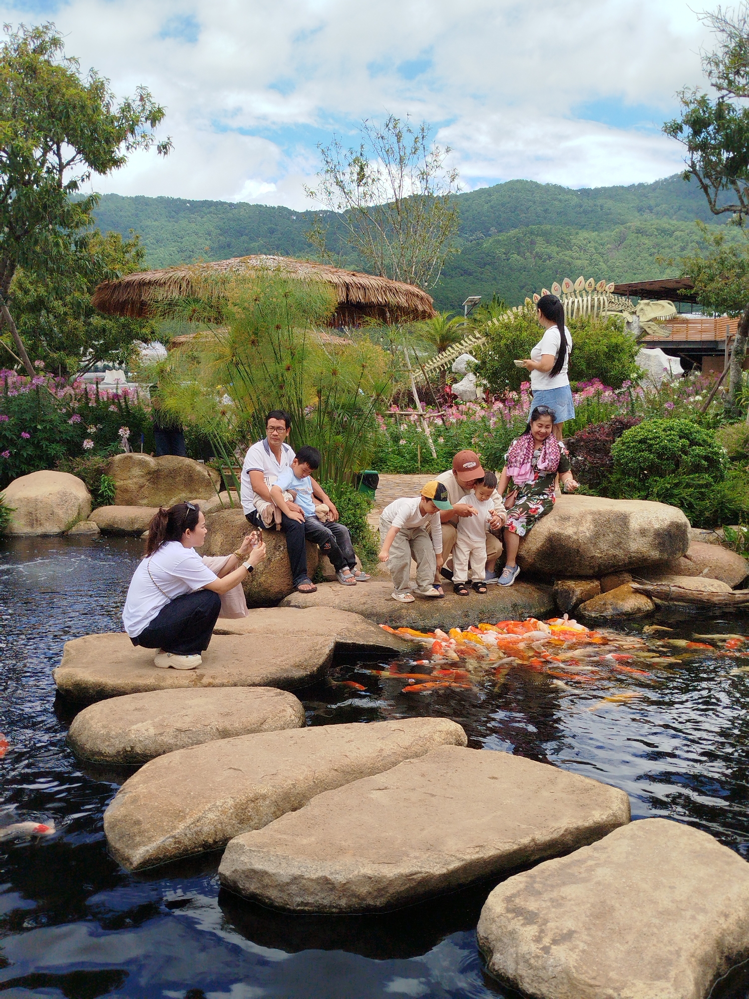 In Lạc Dương, Vietnam, a family is enjoying a day at a park near a pond filled with koi fish. The image shows a large, clear pond with stepping stones leading across it. On the stones and around the pond, several people are gathered.
In the foreground, a woman in black pants and a white t-shirt is crouched on a large stone, taking a photo with her phone. Beside her, on a larger rock, a man in a white polo shirt and dark pants sits with a young boy in a blue shirt on his lap. Another boy in a blue shirt and dark pants is seated next to them.
Further back, on a large rock, a woman in a colorful patterned dress and a pink scarf is seated with two young children. One child, wearing a white shirt and pants, is standing next to her, and another child in a beige outfit is crouched in front of them. A man wearing a brown baseball cap is kneeling next to the children.
In the background, there are lush green hills and trees. A thatched-roof structure is visible, along with a large dinosaur skeleton display and other park elements. The sky is partly cloudy. The koi fish are densely packed in the pond, creating a vibrant spectacle of orange, white, and yellow colors.