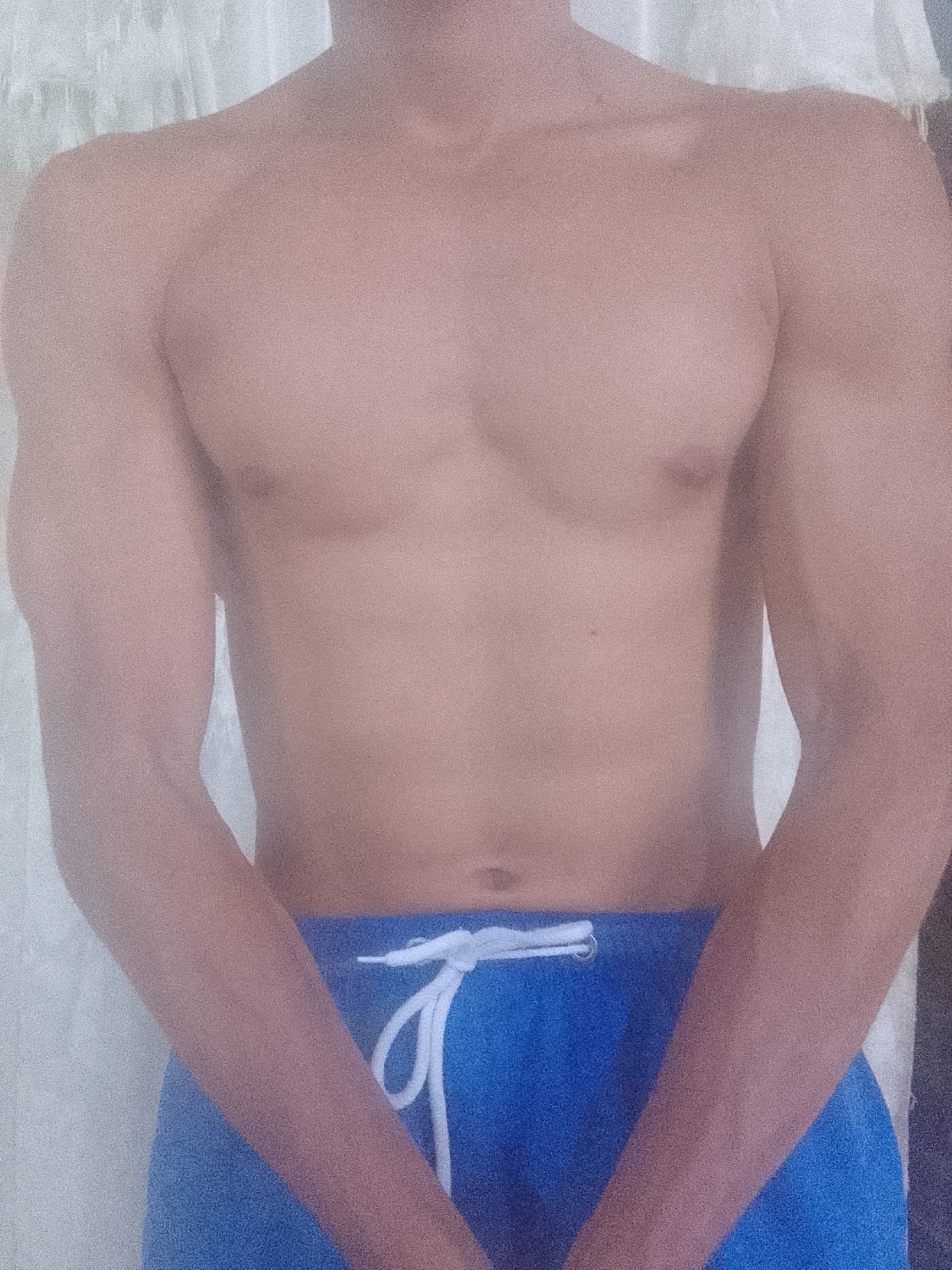 A shirtless person wearing blue shorts stands indoors. The person's hands are folded in front of their abdomen. The location in the city of Juan Griego, Venezuela is not discernible from the image.