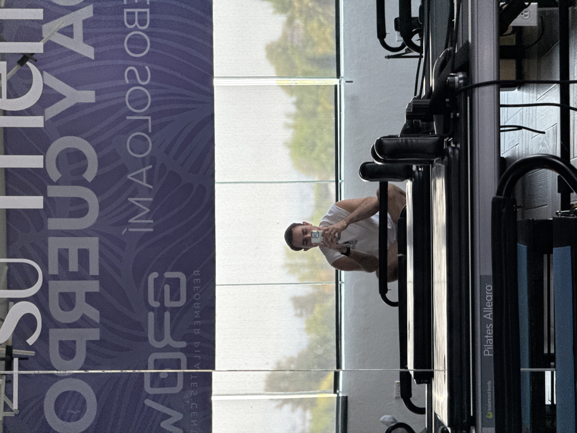 A horizontally-oriented photograph captures a man taking a selfie in a gym setting. The man, positioned in the center of the frame, is holding a cell phone in front of him, presumably taking a picture of himself or the scene. He appears to be on a Pilates Allegro reformer machine. The background reveals a set of large windows looking onto a landscape of trees and greenery, suggesting a natural outdoor setting. To the left of the frame is a banner or sign. The gym appears to be well-lit. The location is unspecified, but given the user's prompt, it is presumed to be located in the city of Puebla, Mexico.