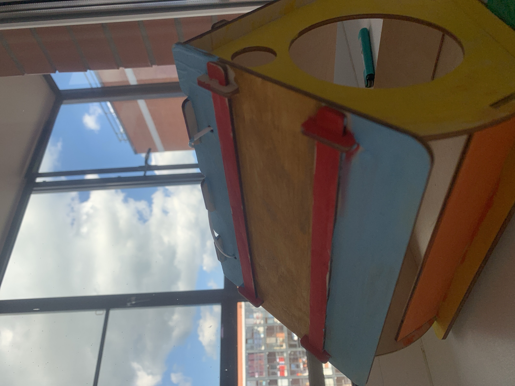 The image shows a close-up, slightly angled view of a wooden craft project, possibly a birdhouse or a model building, painted in various colors including yellow, blue, red, and orange. The craft is positioned in front of a window, with the sky and clouds visible in the background, suggesting it is daytime. Through the window, buildings can be seen, indicating an urban or suburban setting. A teal-colored object, possibly a pen or a lighter, is visible inside a circular opening on the top of the craft. The craft itself is made of thin wooden pieces, assembled together, with some visible joints and cut edges. The paint appears somewhat textured and applied unevenly in places, giving it a handcrafted feel. The lighting suggests natural daylight, with some reflections on the window pane. There is no visible text in the image. The overall impression is of a hobbyist's creation or a child's craft project displayed near a window.