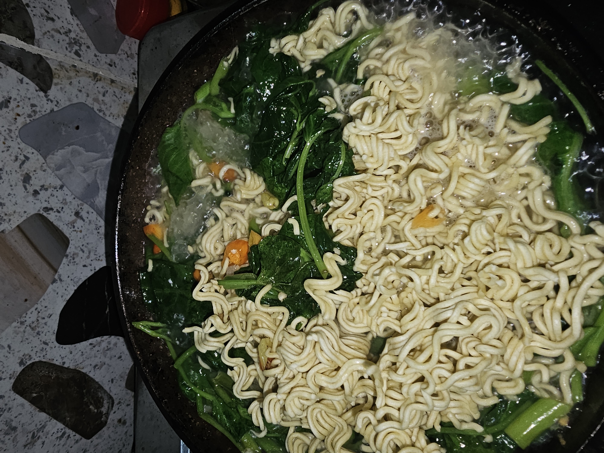This image shows a close-up view of instant noodles being cooked in a dark-colored pan. The noodles are piled high and are interspersed with vibrant green leafy vegetables and small orange pieces, likely chili or other seasoning. The contents of the pan are simmering in a liquid, with some bubbles visible, indicating the cooking process. The pan is set against a background that appears to be a countertop with a speckled, mosaic-like pattern in shades of gray, white, and black. There are also dark, amorphous shapes within the pattern. In the upper right corner, a small red object, possibly a lid or a container, is partially visible. The overall impression is of a home-cooked meal being prepared. While the image depicts a common culinary scene, there is no information within the image itself to pinpoint its location to Lamongan, Indonesia.