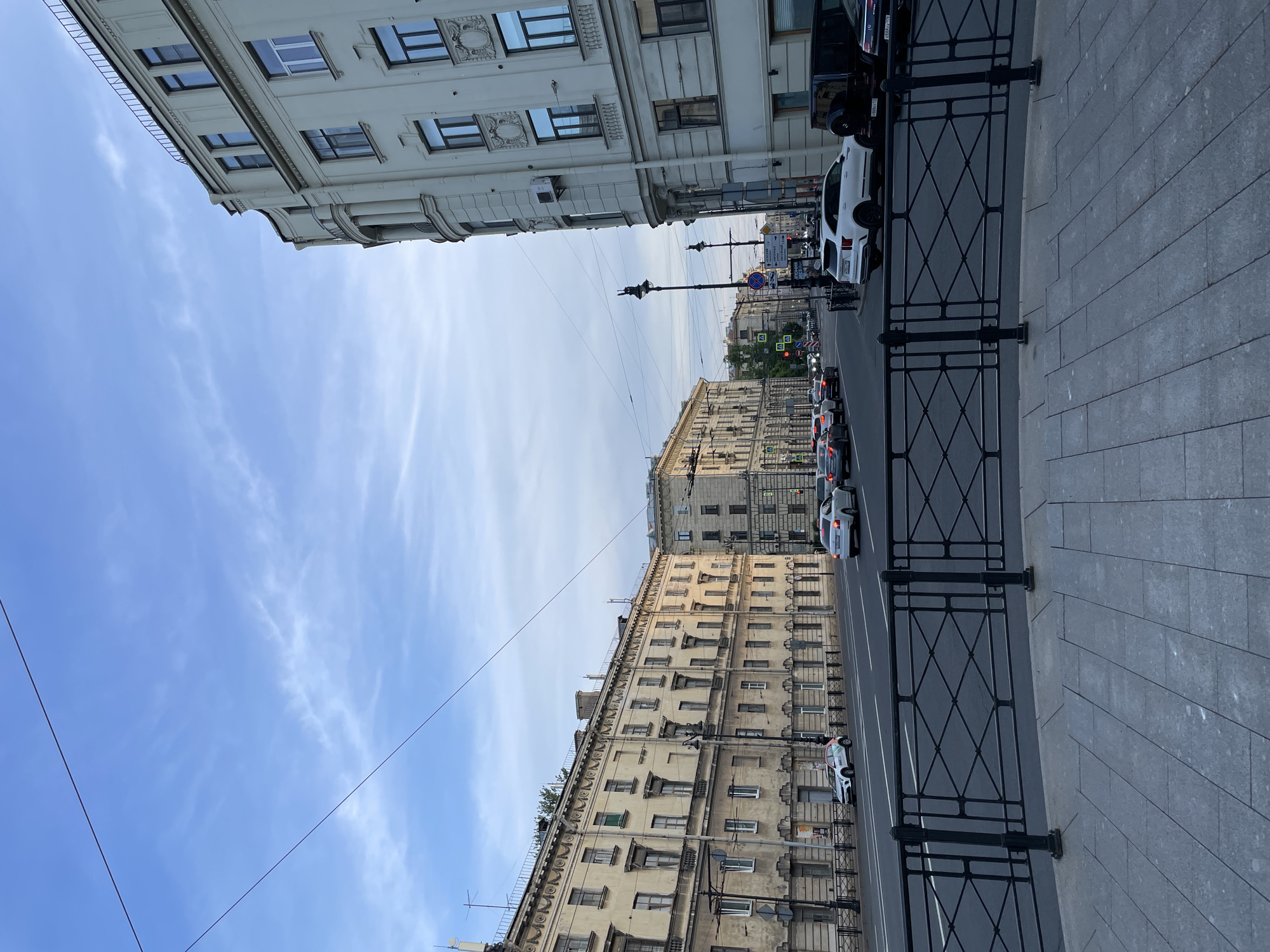 The image displays a street view in Saint Petersburg, Russia, looking south along Ligovsky Prospekt, likely from the area of Ploshchad Vosstaniya. In the immediate foreground, a dark gray paved pedestrian walkway with rectangular blocks is bordered by a curved black metal railing featuring diamond-patterned panels.
Beyond the railing lies a multi-lane asphalt road with white lane markings. On the left side of the road, a long, multi-story historical building with a pale yellow, somewhat weathered facade extends into the distance. This building features numerous rectangular windows, many appearing dark, and decorative elements such as cornices and textured panels. A white car, identified by "Делимобиль" (Delimobil, a car-sharing service) branding, is parked facing the viewer. Further down, more buildings and trees are visible. Classic black ornate street lamps are present along this side of the street.
On the right side of the road, a lighter cream-colored, multi-story building with a prominent rounded corner dominates the mid-ground. This building exhibits intricate architectural details, including a curved balcony with columns on an upper floor and decorative panels between its large, dark windows. An air conditioning unit is mounted on its side. Several vehicles are parked along the curb: a white SUV (likely a Kia Sorento), a black Mercedes G-Wagen, and a dark sedan. Overhead wires traverse the sky. Road signs featuring Cyrillic script are visible, with prominent mentions of "Лиговский проспект" (Ligovsky Prospekt) and "Невский проспект" (Nevsky Prospekt). The sky above is light blue with scattered, wispy white clouds. No individual people are distinctly visible or engaged in specific actions within the frame.