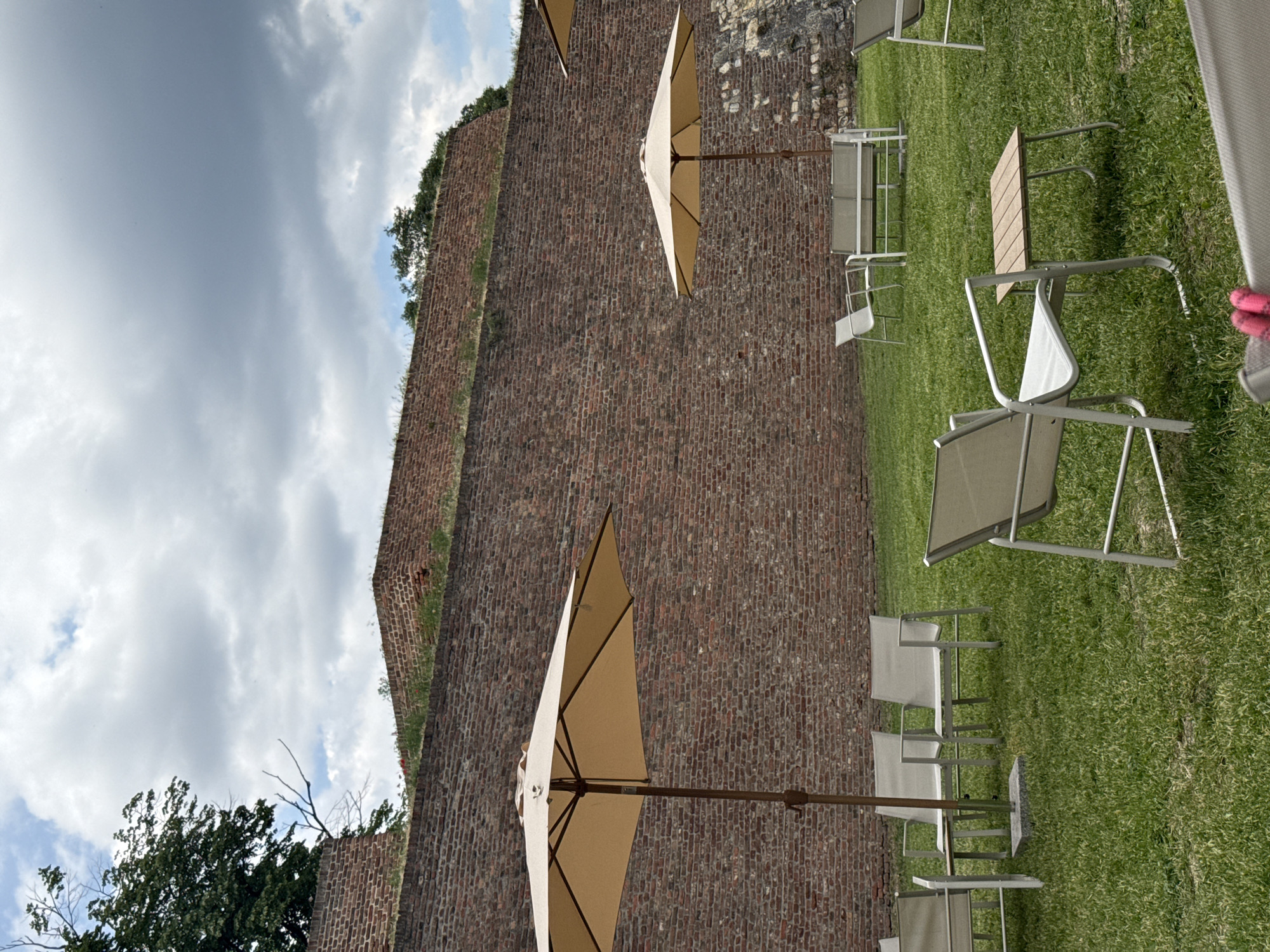 The scene shows an outdoor seating area with multiple chairs and tables on a lawn near a brick wall. There are two tan umbrellas in the image. The brick wall could be the one in Belgrade Fortress. The sky has cloudy weather.
