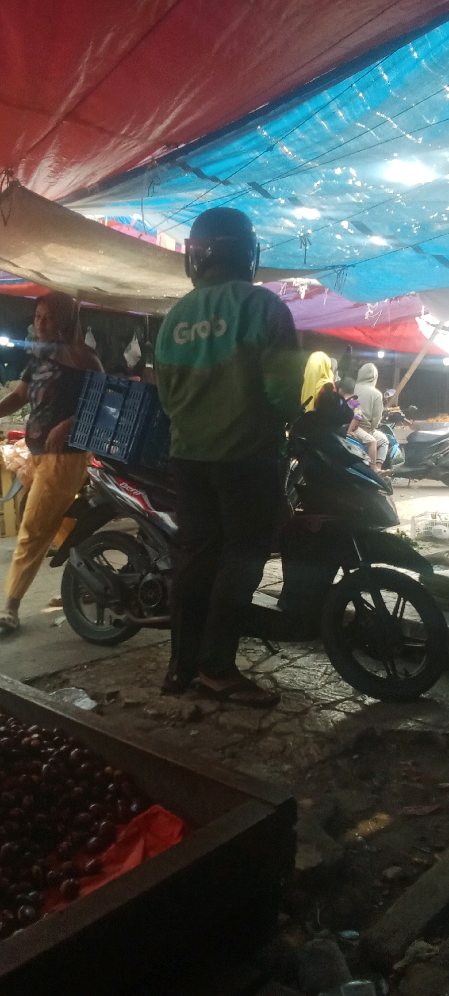 The image depicts a street scene, possibly a market, under a makeshift canopy of tarps in Karawang, Indonesia. A person wearing a dark helmet and a green jacket with the "Grab" logo is standing next to a motorcycle. The jacket has two horizontal bands, a lighter green on top and a darker green on the bottom, with the "Grab" logo in white on the upper band. The motorcycle is dark-colored and appears to be a scooter or an underbone style. In the background, another person in a dark t-shirt and yellow pants is visible, holding a blue plastic crate. Further back, several other individuals are present, some sitting on motorcycles. The ground is paved and appears wet or reflective in places. In the foreground, a wooden bin filled with dark, round objects, possibly fruit, is visible. The overall lighting suggests it is either dusk or a dimly lit covered area.