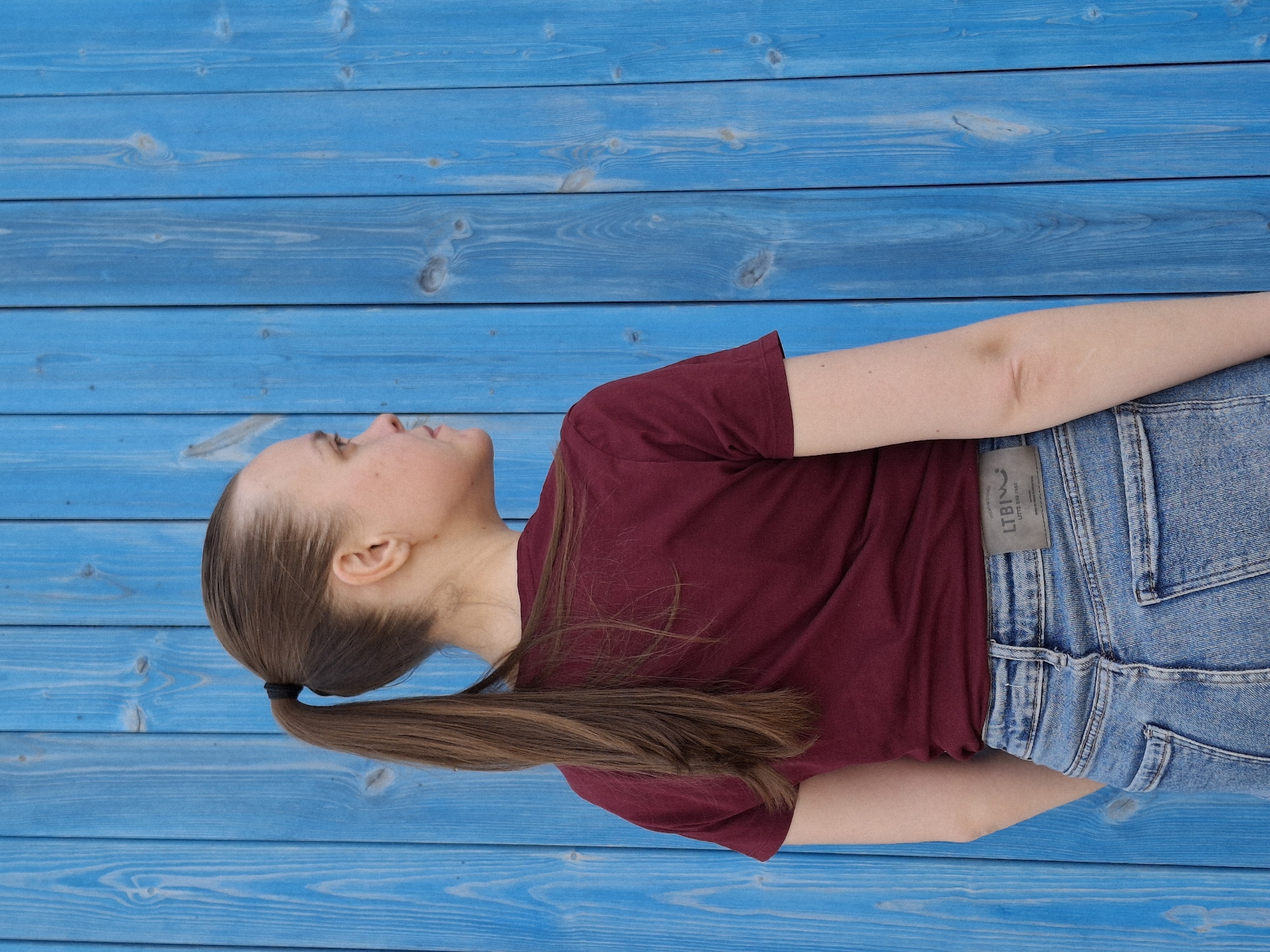 A young woman lies on her side on a blue wooden surface, possibly a deck or platform. She is wearing a burgundy t-shirt and blue jeans with a visible LTBI tag. Her brown hair is pulled back into a ponytail. The background consists entirely of the blue wooden surface. The location is Chaikovsky, Russia.